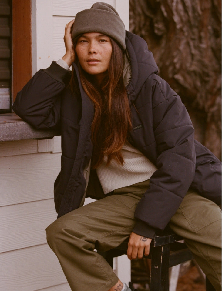Adventure-ready woman with brown hair wearing a winter parka with a hood in a rugged outdoor setting, embodying the spirit of Roark's travel and adventure essentials.