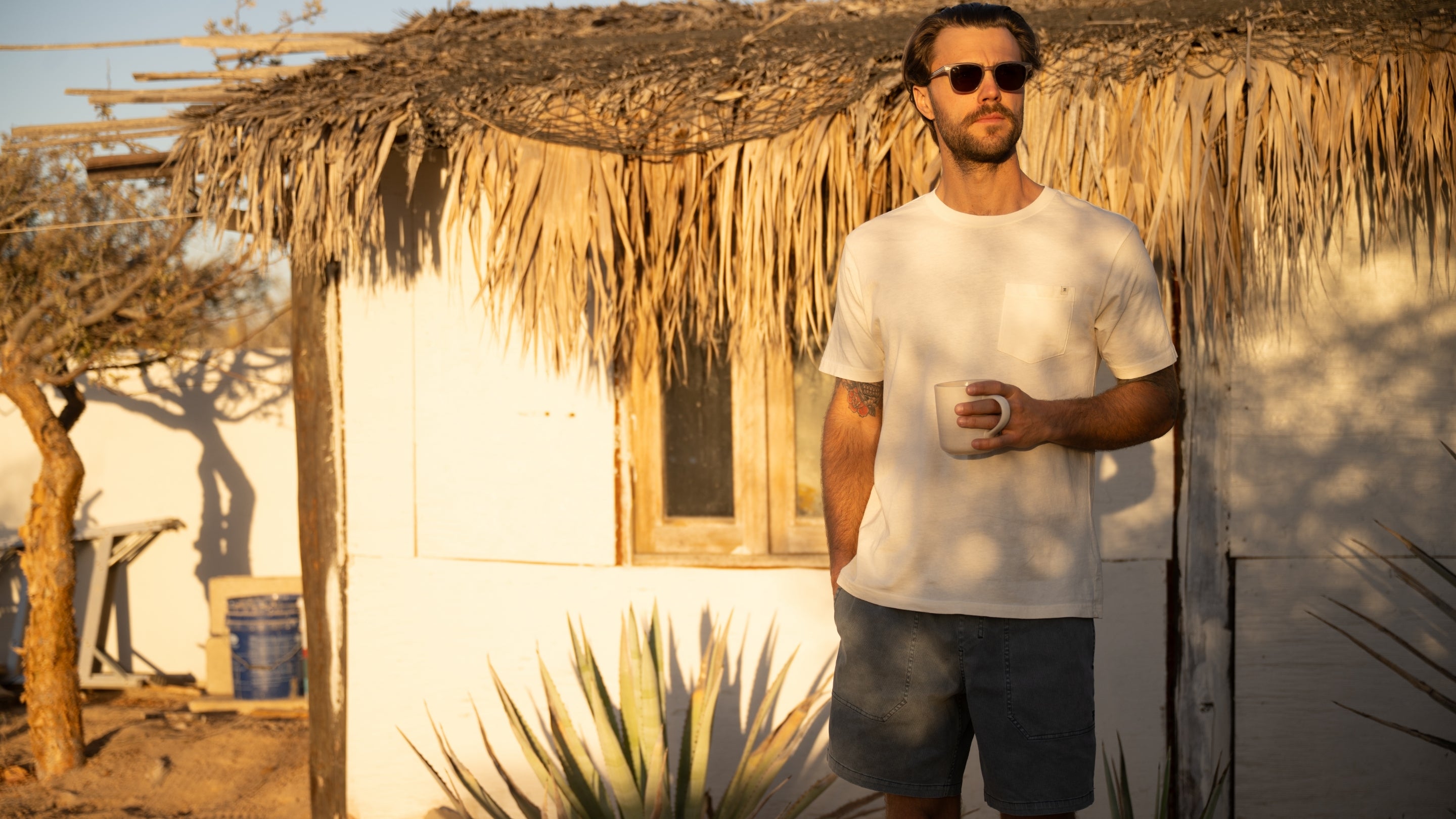 Bearded man standing in a summer setting wearing a basic Roark T-shirt, embodying the adventure and travel spirit