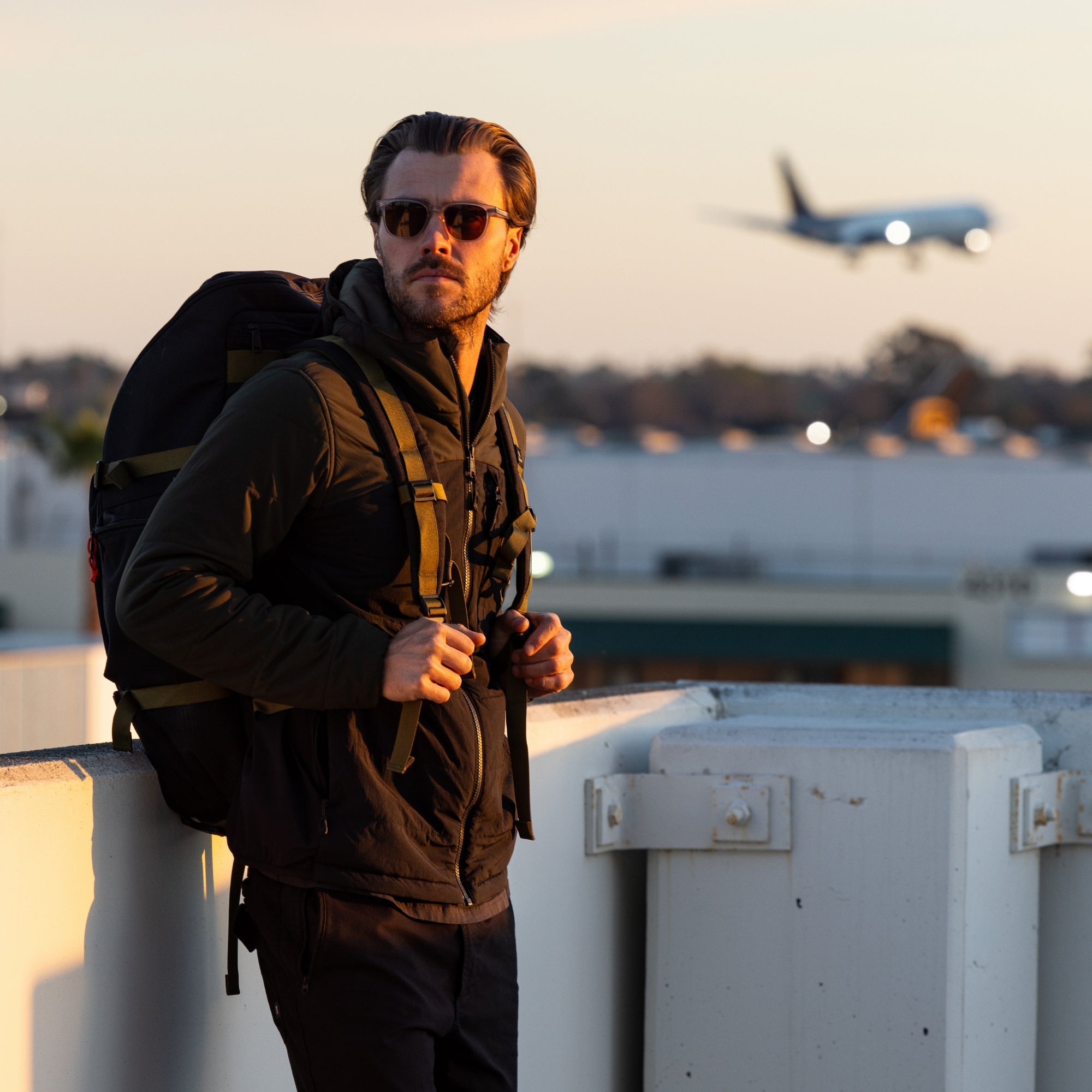 Stylish man in sunglasses wearing an olive jacket with a backpack, standing outdoors during sunset with an airplane in the background.