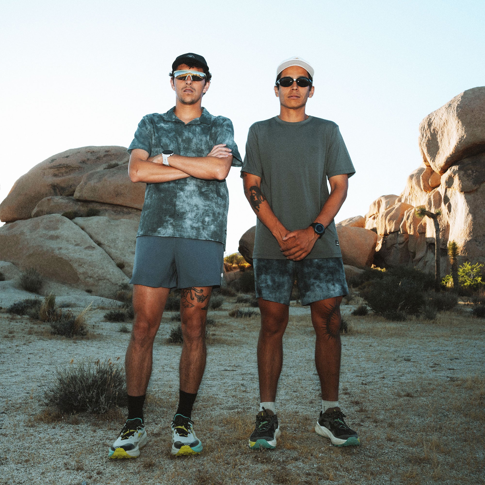 Two men wearing teal tie-dye short-sleeve tees and shorts stand in a desert landscape with large boulder formations