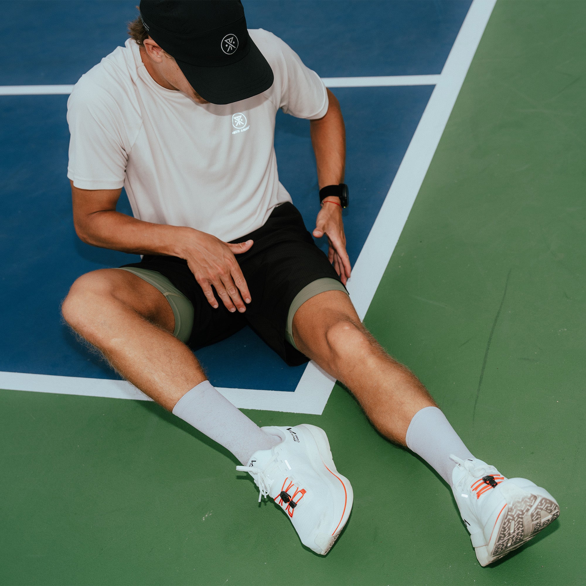 Tennis player wearing white Alta Elite Trail short sleeve tee with black cap and shorts on blue and green court