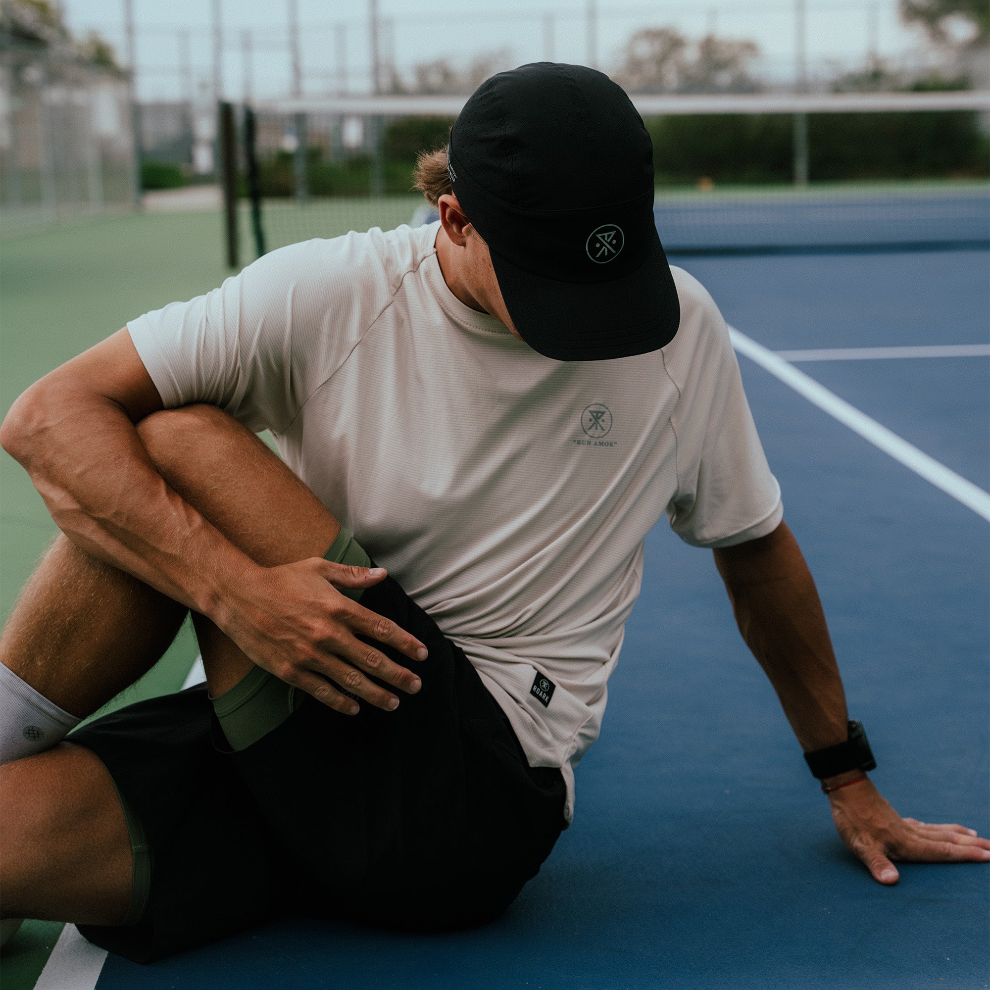 Man wearing beige Alta Elite Trail short sleeve tee and black cap sitting on blue tennis court