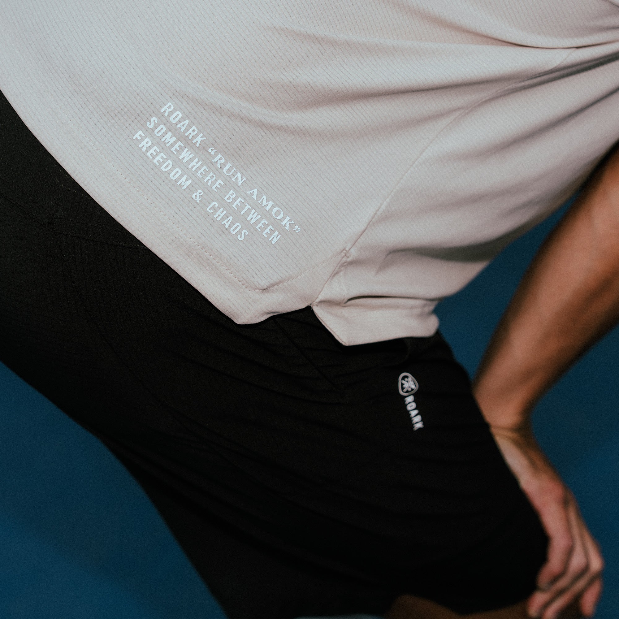 White short-sleeve tech tee with roark grown text and logo worn with black shorts against blue background