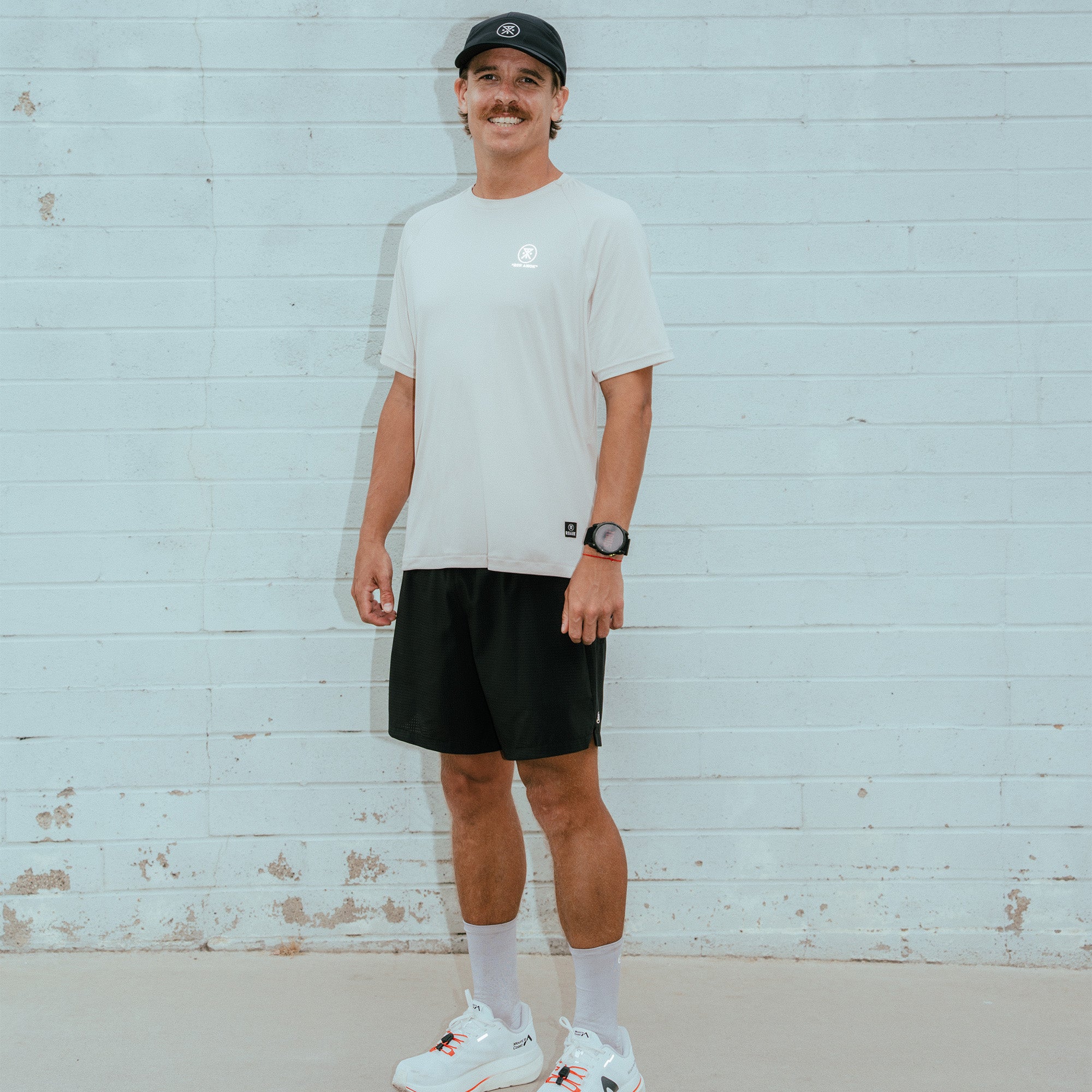 Man wearing white short-sleeve t-shirt with small logo and black shorts standing against light blue wooden wall