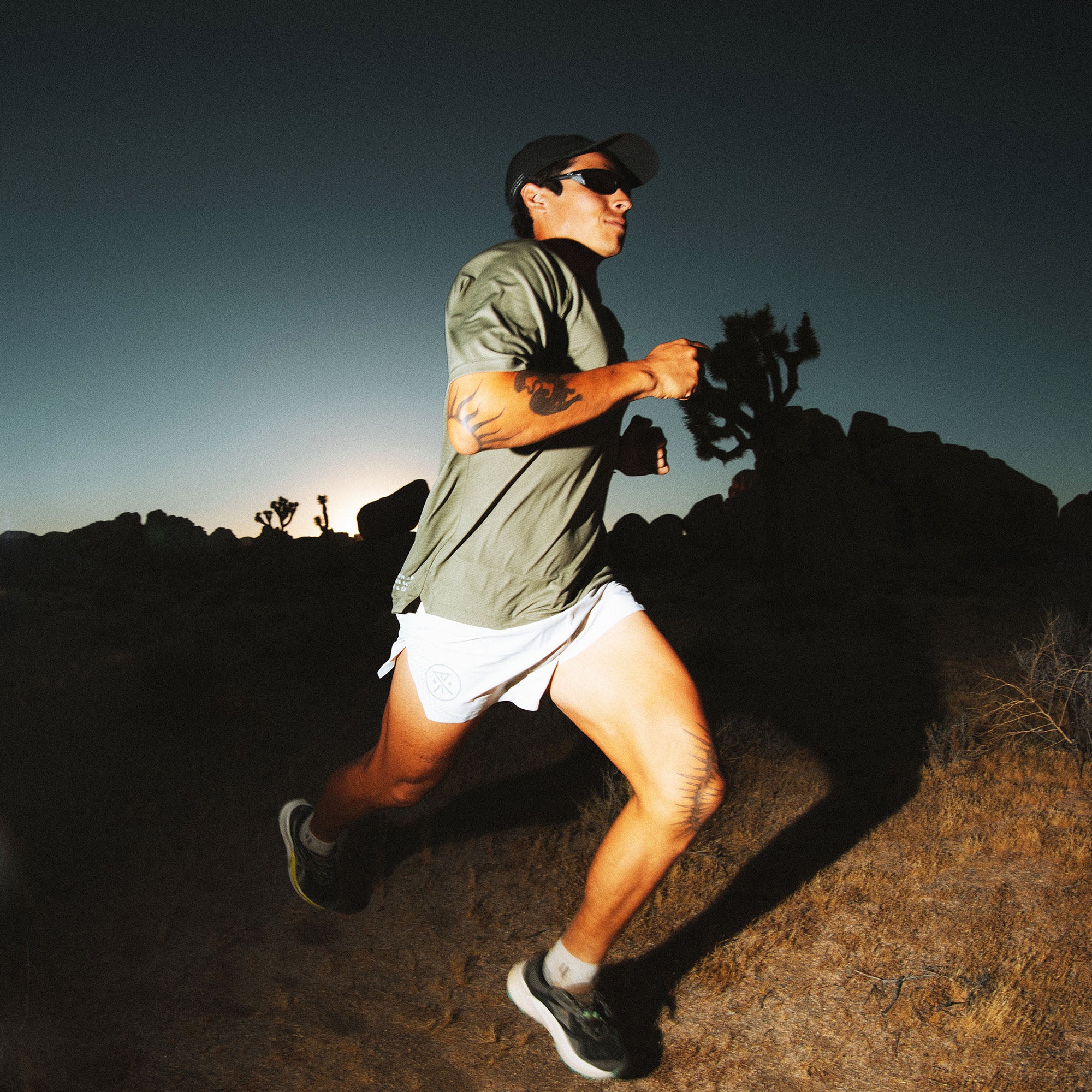 Man trail running in olive green short-sleeve tee, white shorts, and black sneakers on desert terrain at dusk