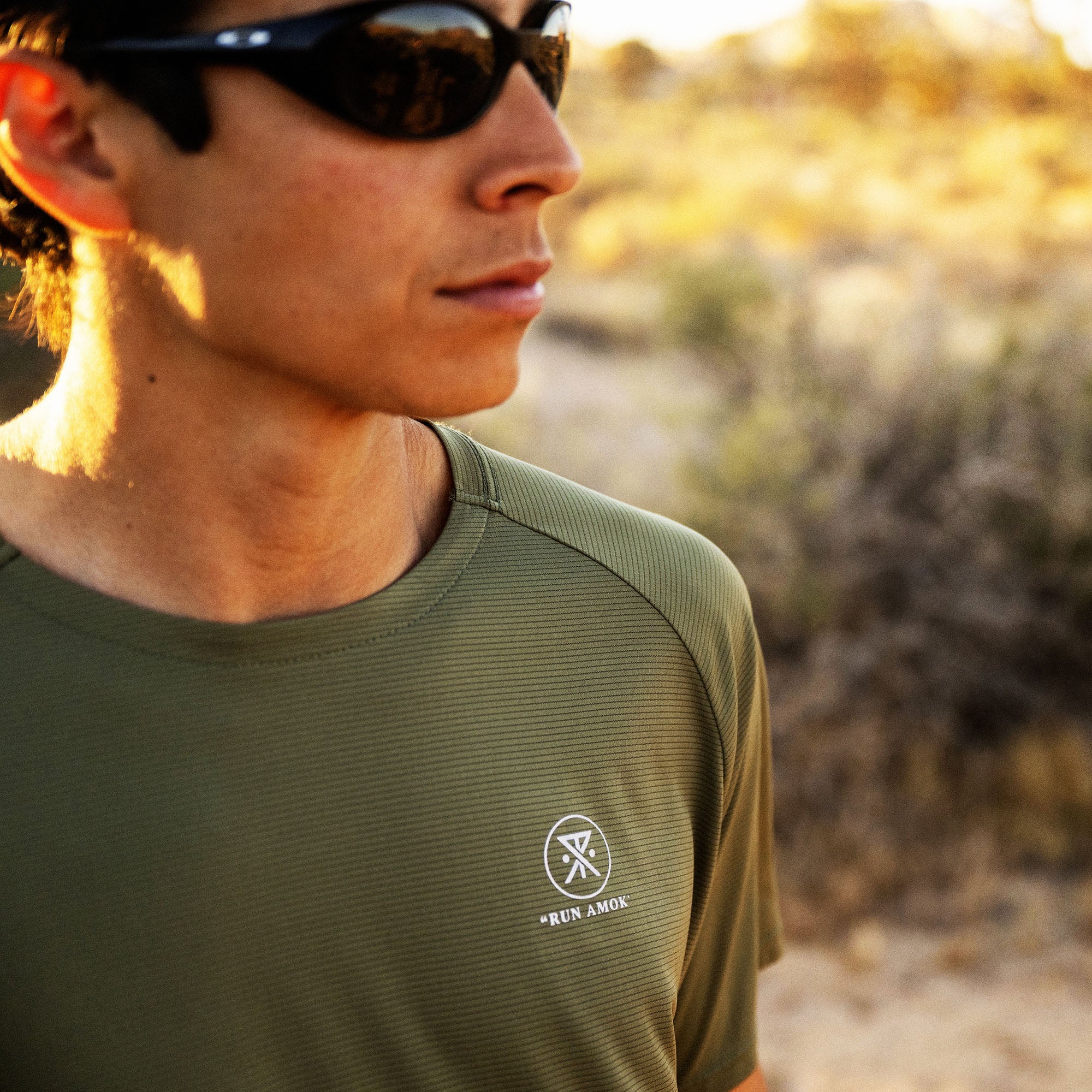 Man wearing olive green Alta Elite Trail short sleeve tee with Run Amok logo, viewed from side in desert landscape