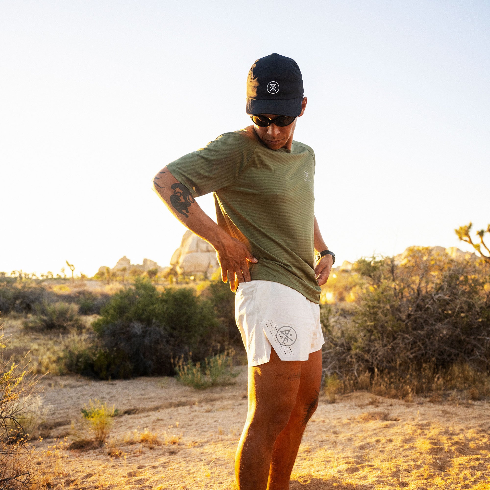 Man wearing olive green short-sleeve tee and white athletic shorts while running in desert landscape at sunset