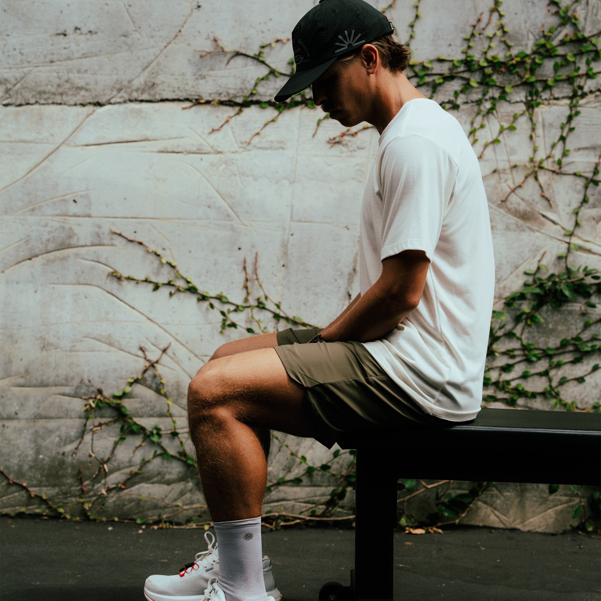 Man wearing white Alta Elite Trail short sleeve tee and khaki shorts, sitting on bench against ivy-covered concrete wall
