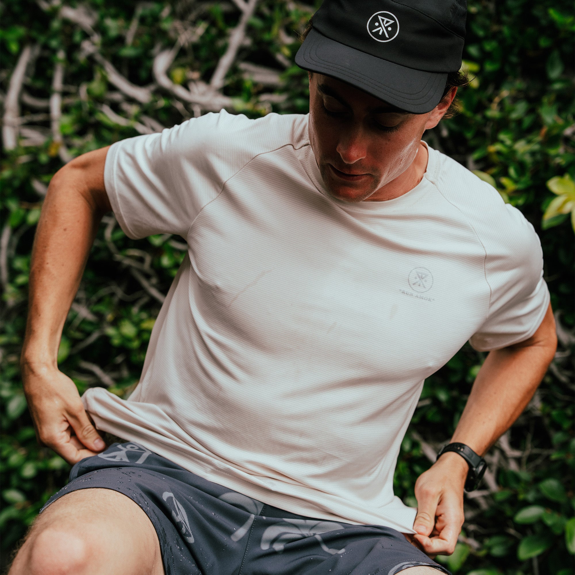Man wearing cream-colored short-sleeve athletic tee with embroidered logo, paired with black cap and dark shorts outdoors