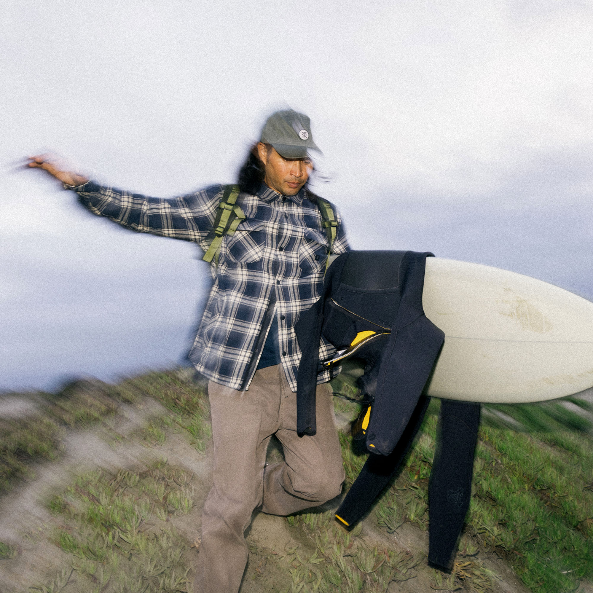 Outdoor enthusiast wearing a Roark Backwoods Overshirt Jacket in flannel, styled with a baseball cap, ready for a surfing adventure.