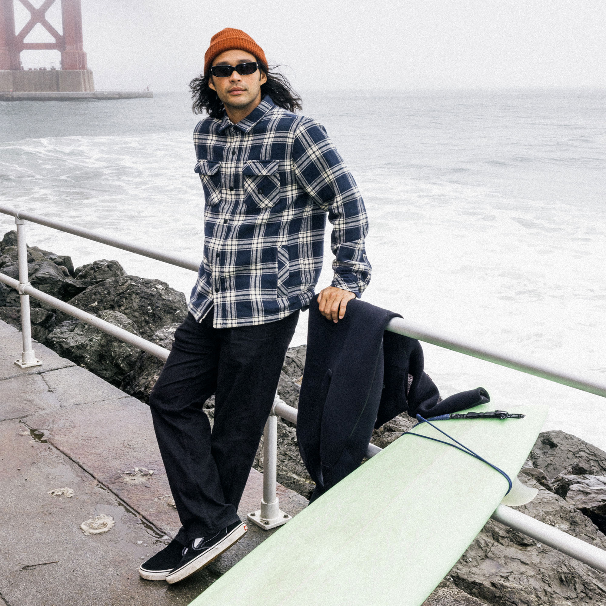Model showcasing outdoor style in a flannel Backwoods Overshirt Jacket by Roark, paired with casual trousers and rugged shoes, standing adventurous in a natural setting.