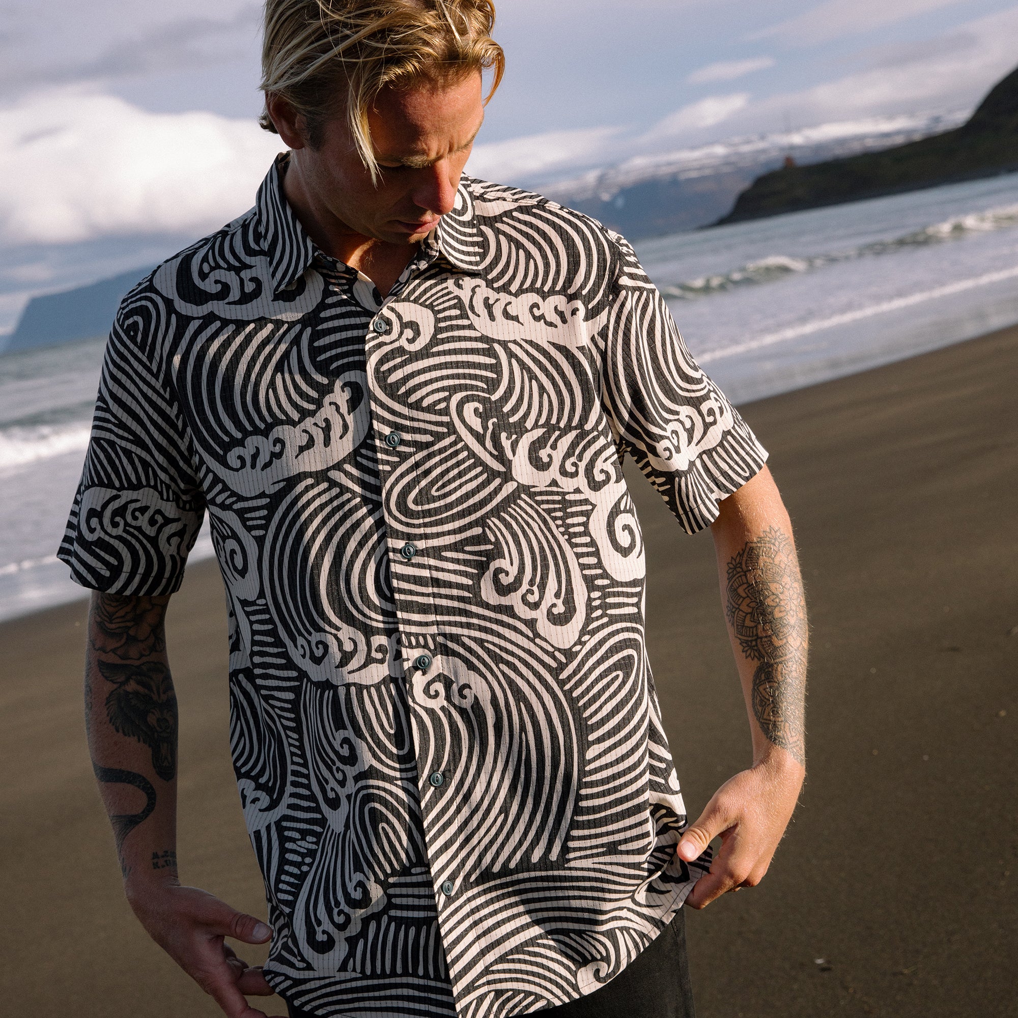 Black and white wave patterned short sleeve shirt worn by man on beach with ocean and mountains in background