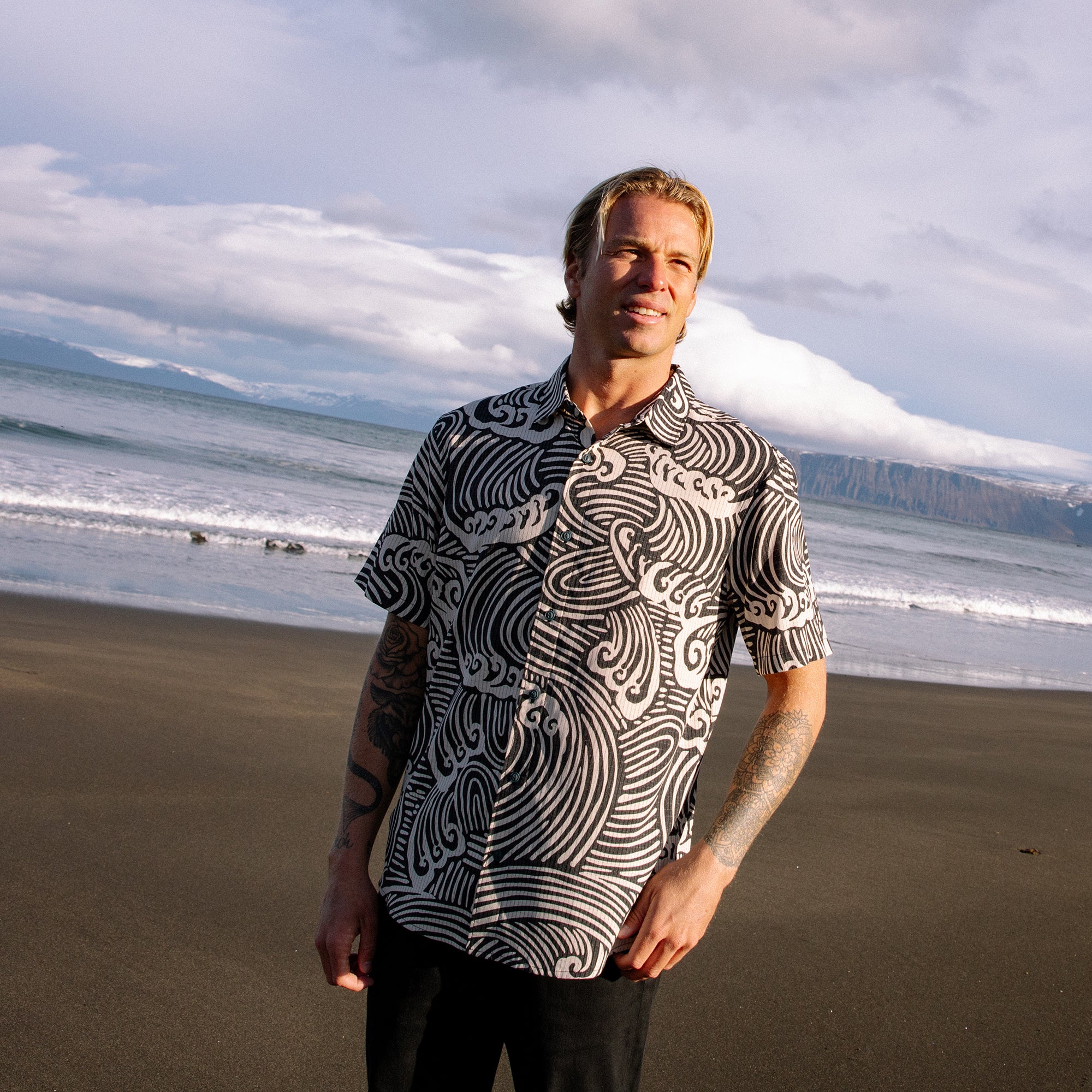 Man wearing black and white wave patterned short sleeve shirt standing on beach with ocean and mountains in background