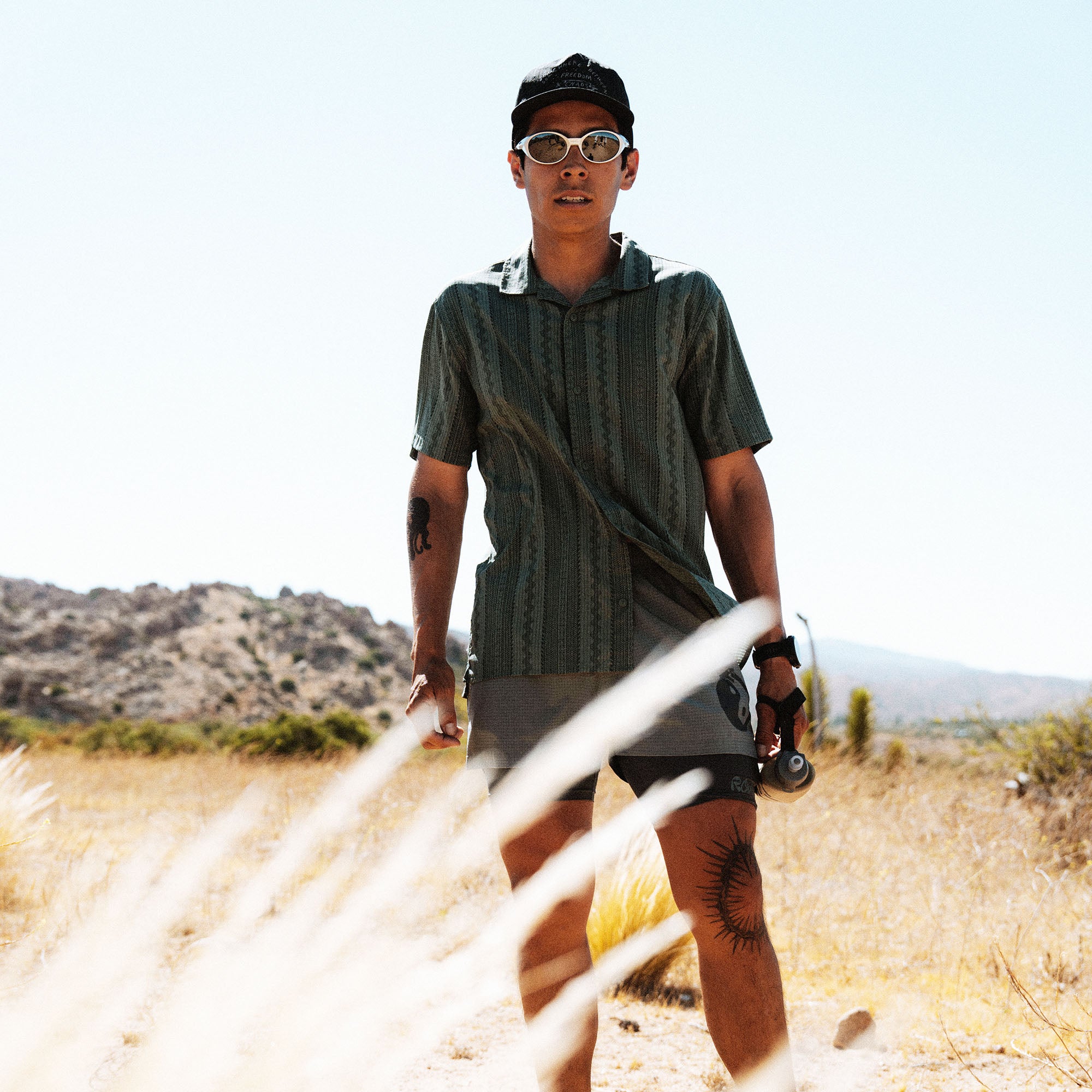 Man wearing olive green striped shirt and sunglasses holding skateboard in desert landscape with rocky hills