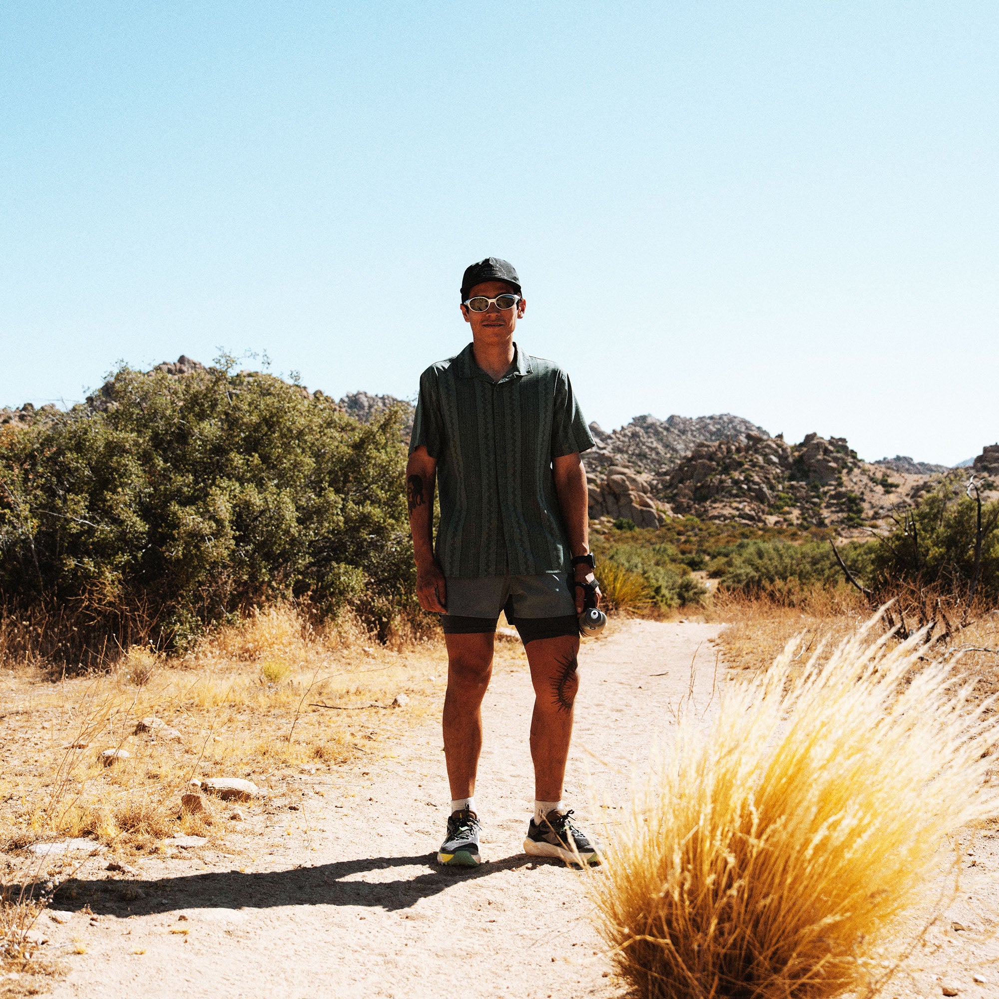 Male runner in dark short-sleeve shirt and black shorts jogging on desert trail with rocky hills and desert vegetation under clear sky