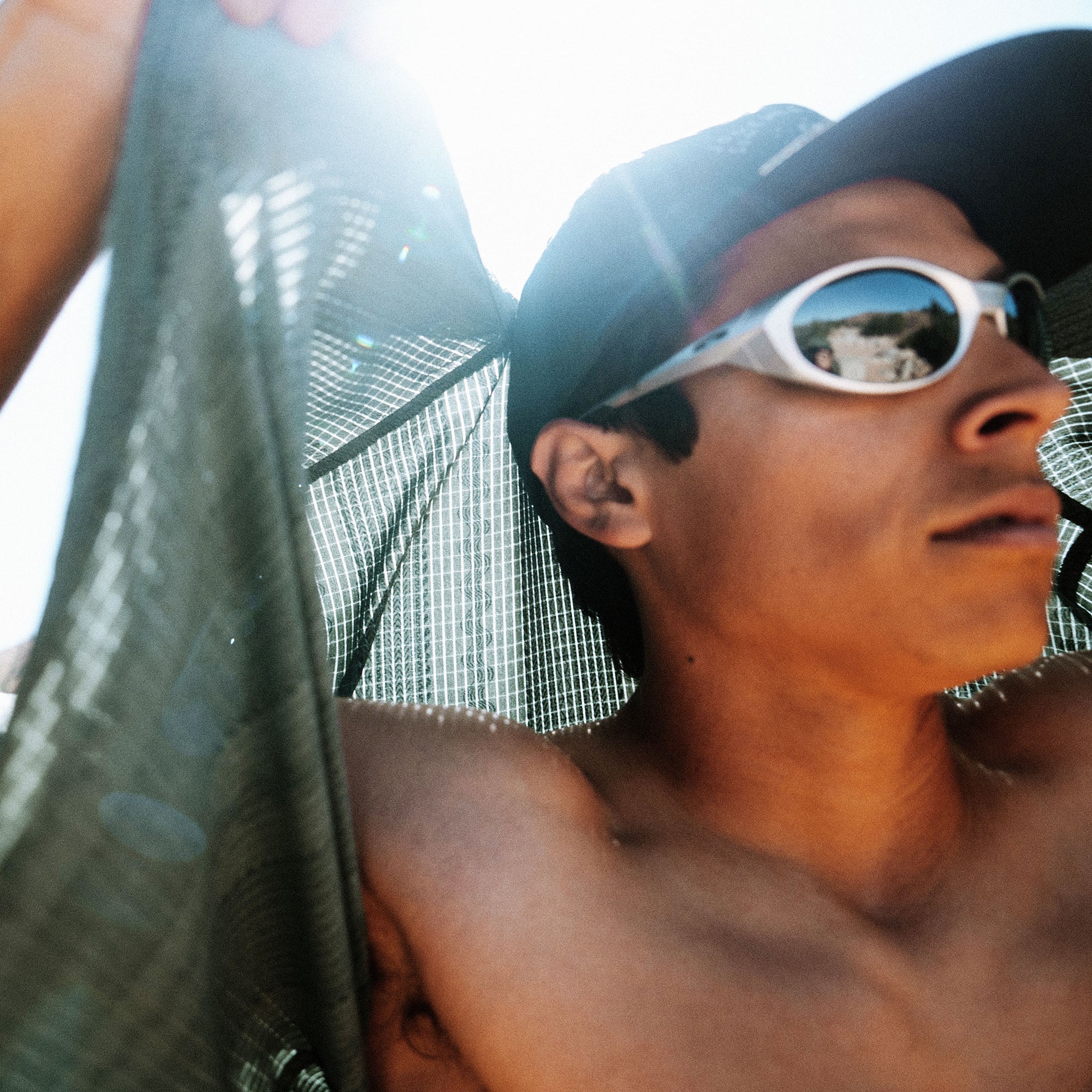 Person wearing white oval sunglasses and black baseball cap with mesh panels, holding green fabric outdoors in bright sunlight