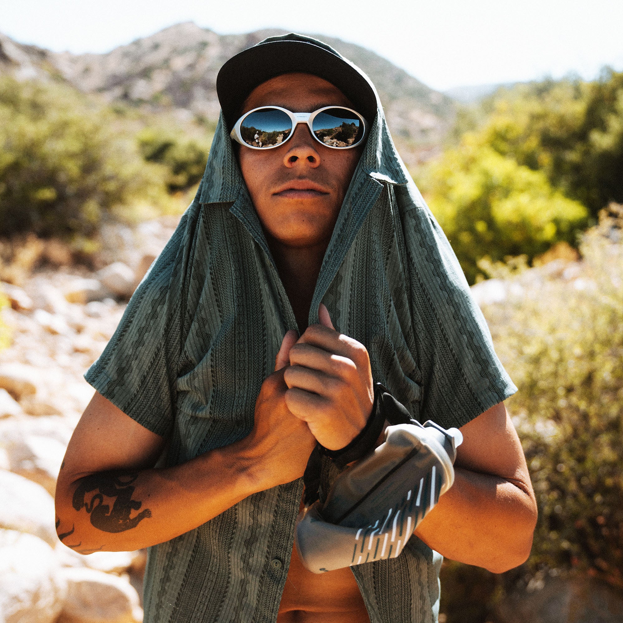 Person wearing white oval sunglasses and black cap with teal gauze head wrap in desert mountain landscape