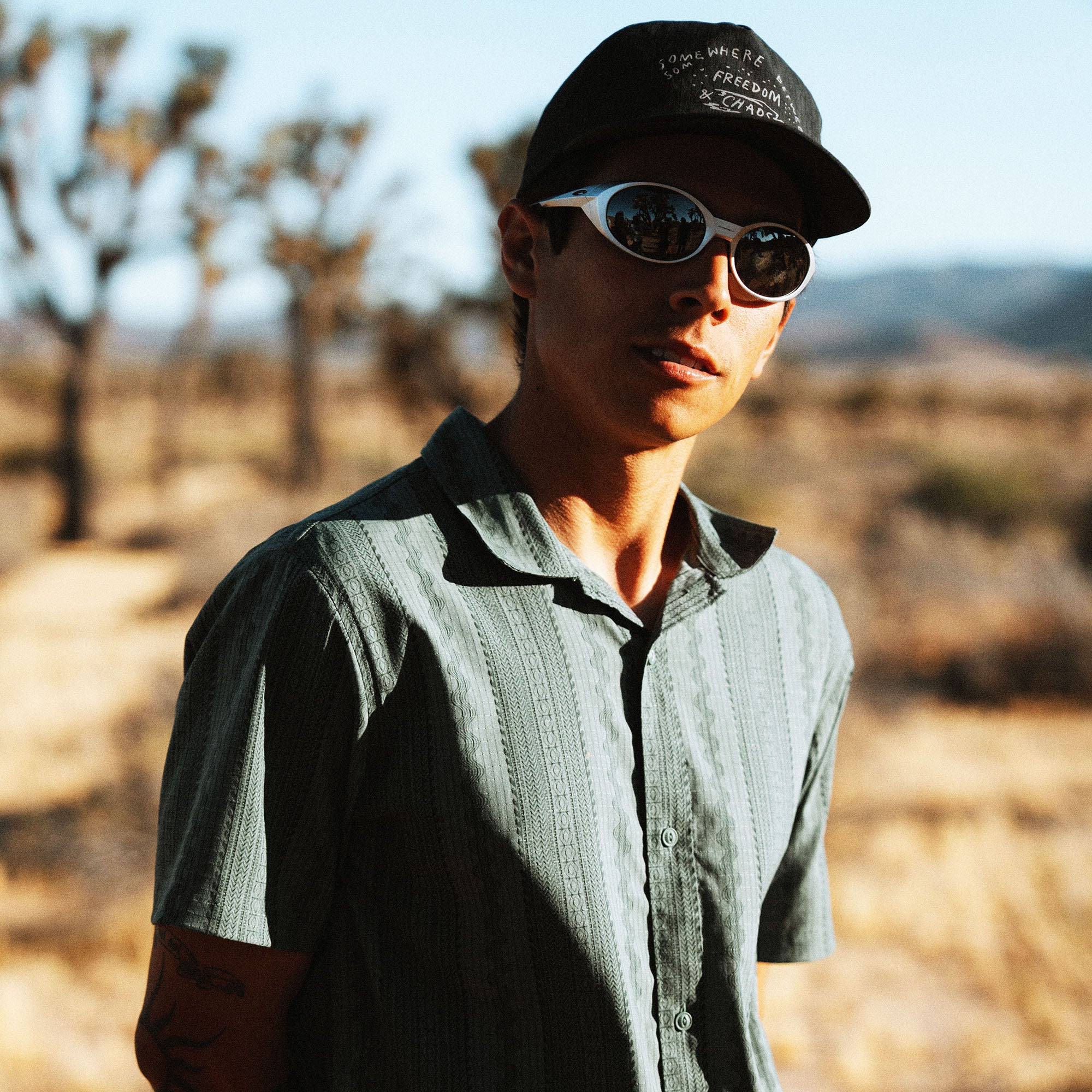 Man wearing black cap and oval sunglasses with light blue shirt in desert landscape with Joshua trees