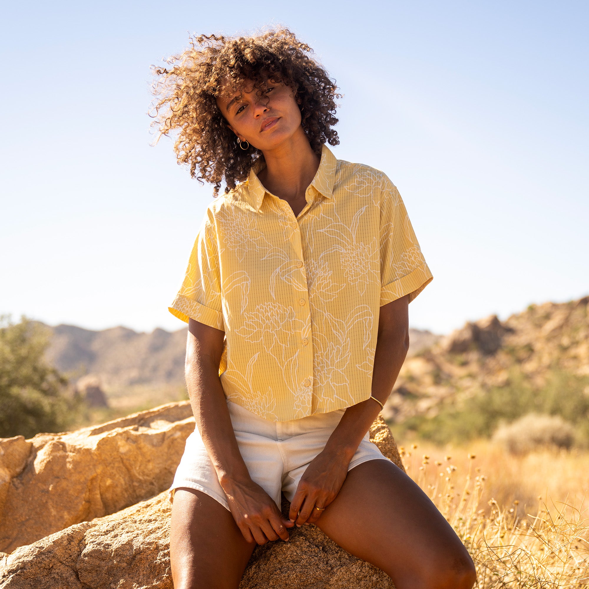 Woman wearing yellow short-sleeve button-up shirt with white leaf print and white shorts, sitting outdoors in desert landscape