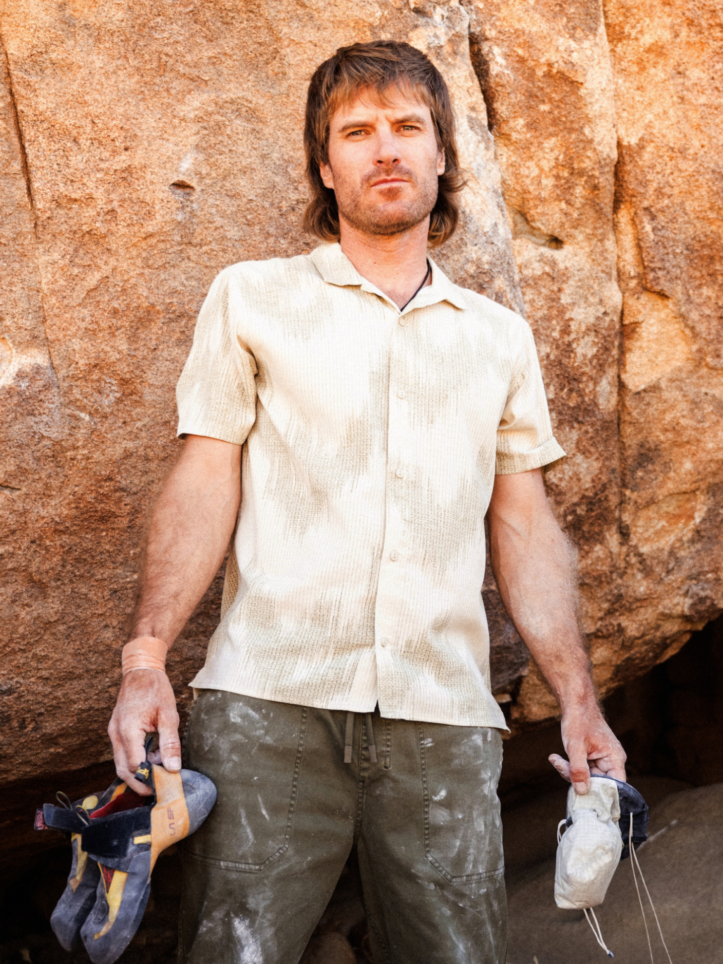 Man wearing cream textured short-sleeve shirt and olive work pants, holding work gloves against weathered brick wall