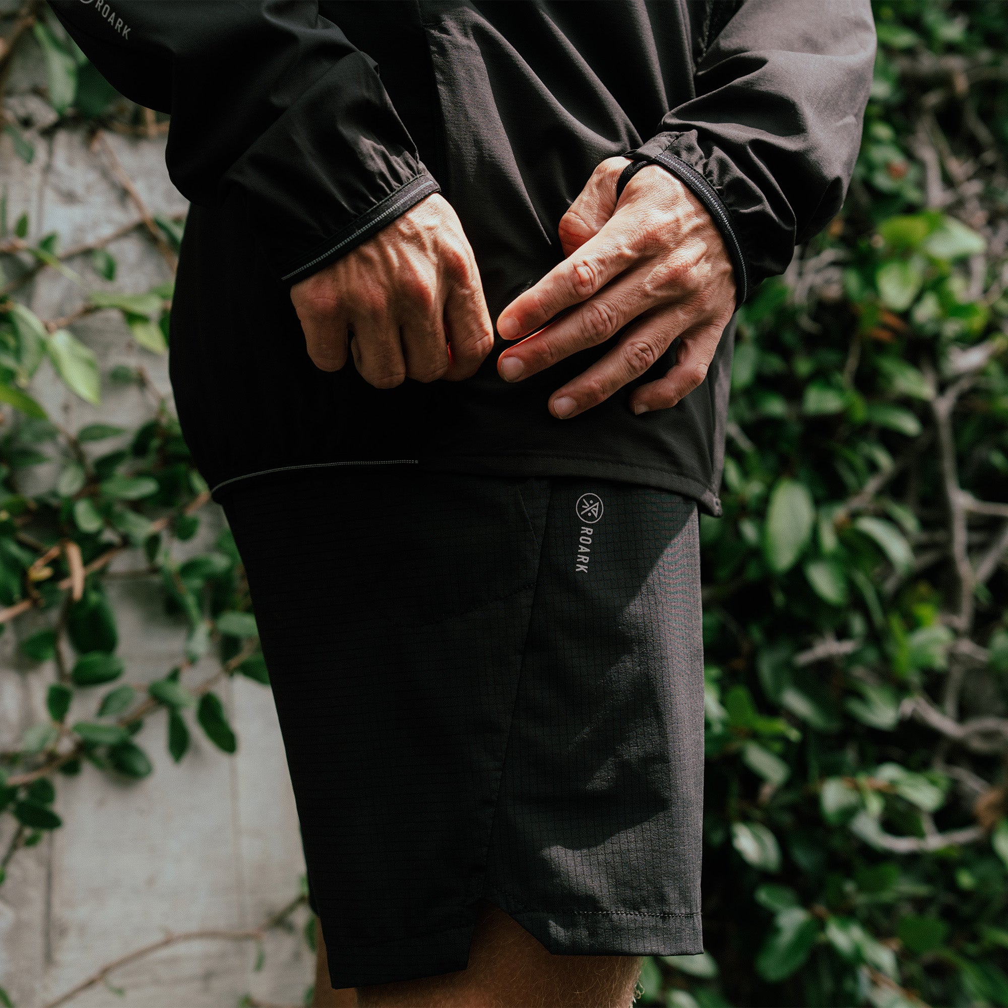 Black nylon shorts with textured inseam detail, worn with matching jacket, hands in pockets against green foliage background