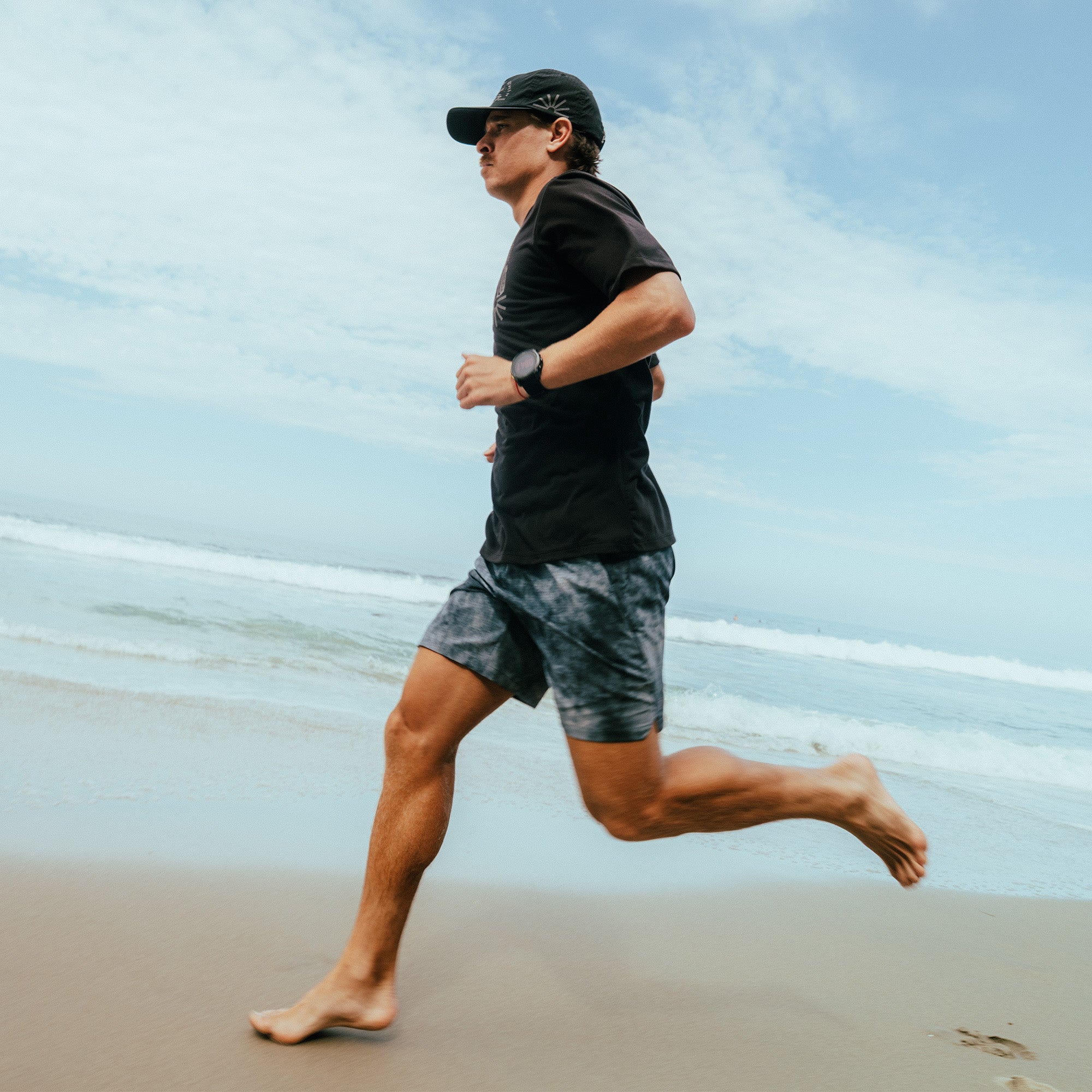 Man running on beach wearing dark slate Bommer shorts, black t-shirt, and baseball cap with ocean and sky in background