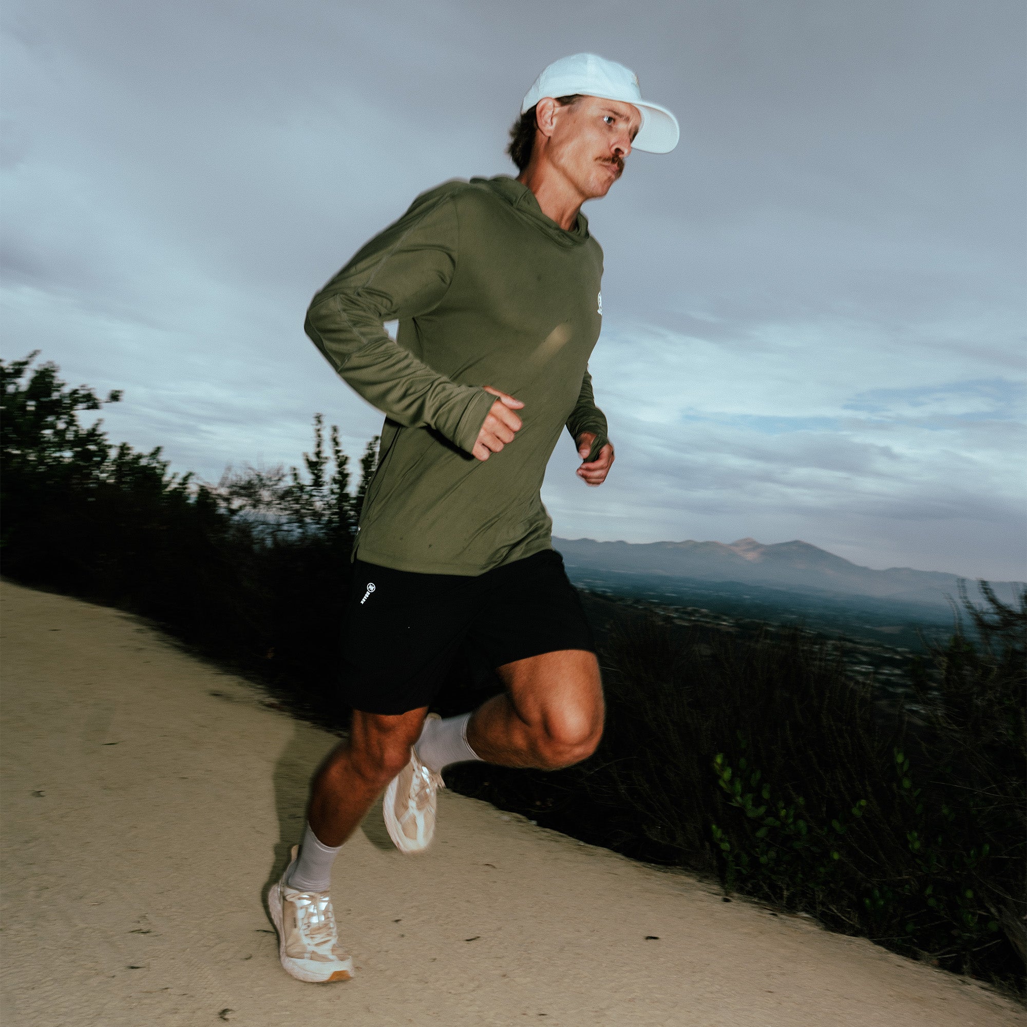 Man jogging outdoors in black Bommer shorts with olive long-sleeve shirt and white cap on mountain trail