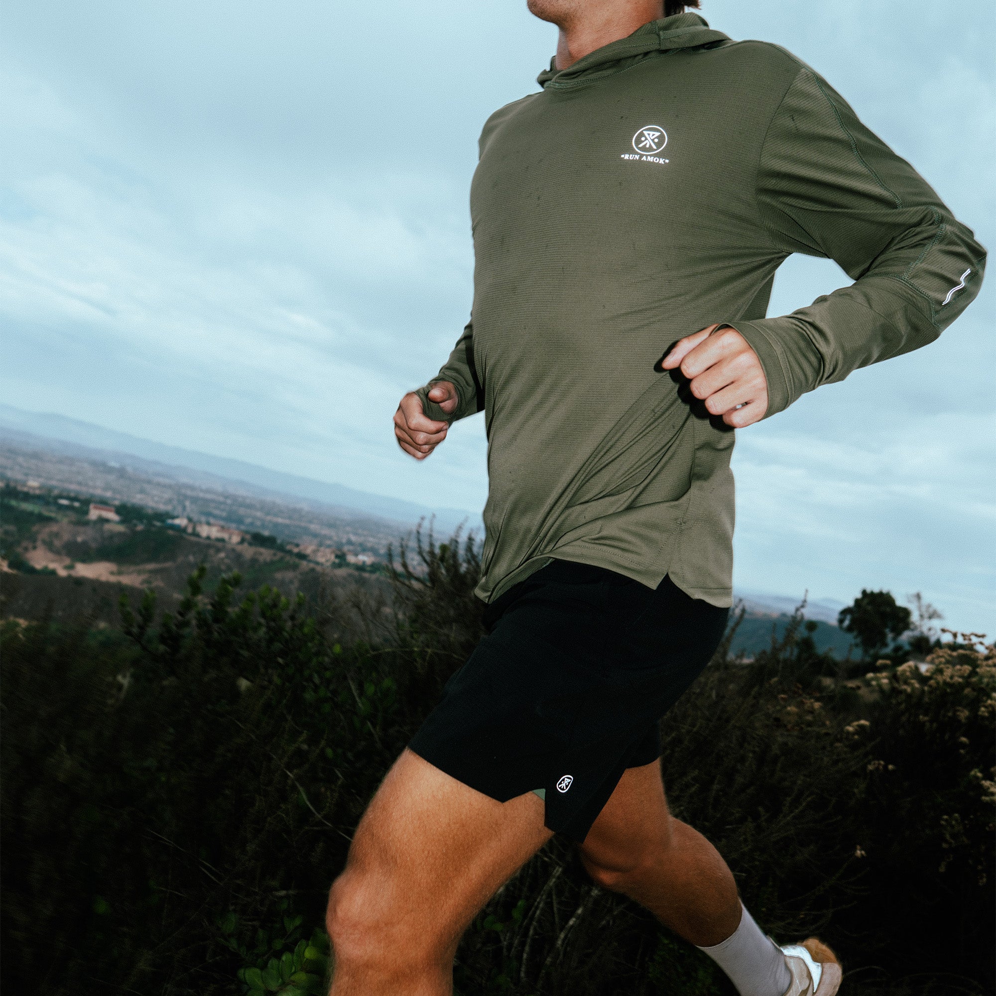 Man jogging outdoors in olive green hoodie and black shorts on a hillside with valley landscape view