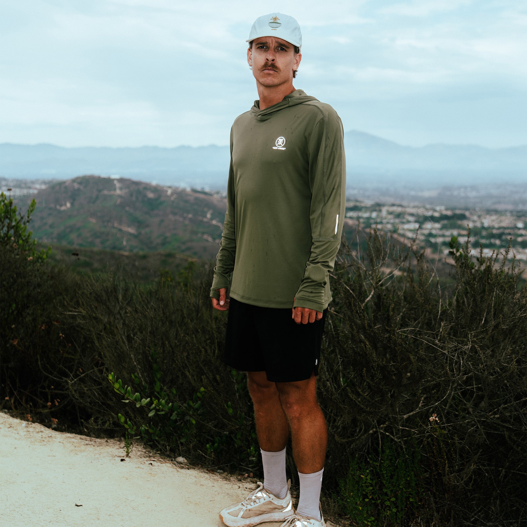 Man wearing olive green hoodie with small white logo, black shorts, light blue cap, and white socks standing on hillside trail overlooking