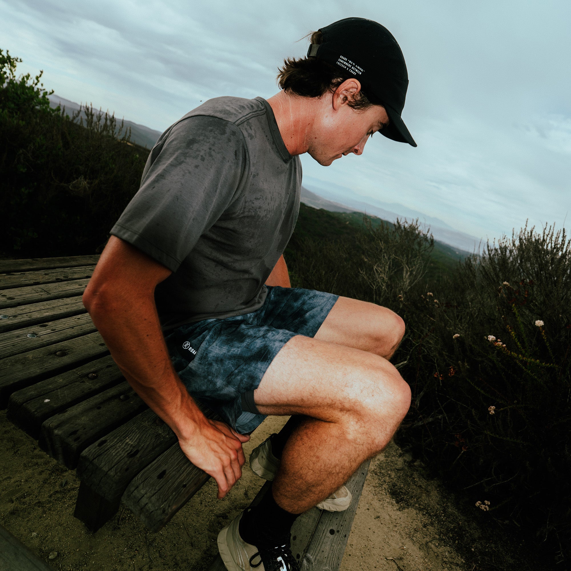 Man wearing charcoal t-shirt and blue patterned shorts sitting outdoors on wooden bench with mountain landscape background