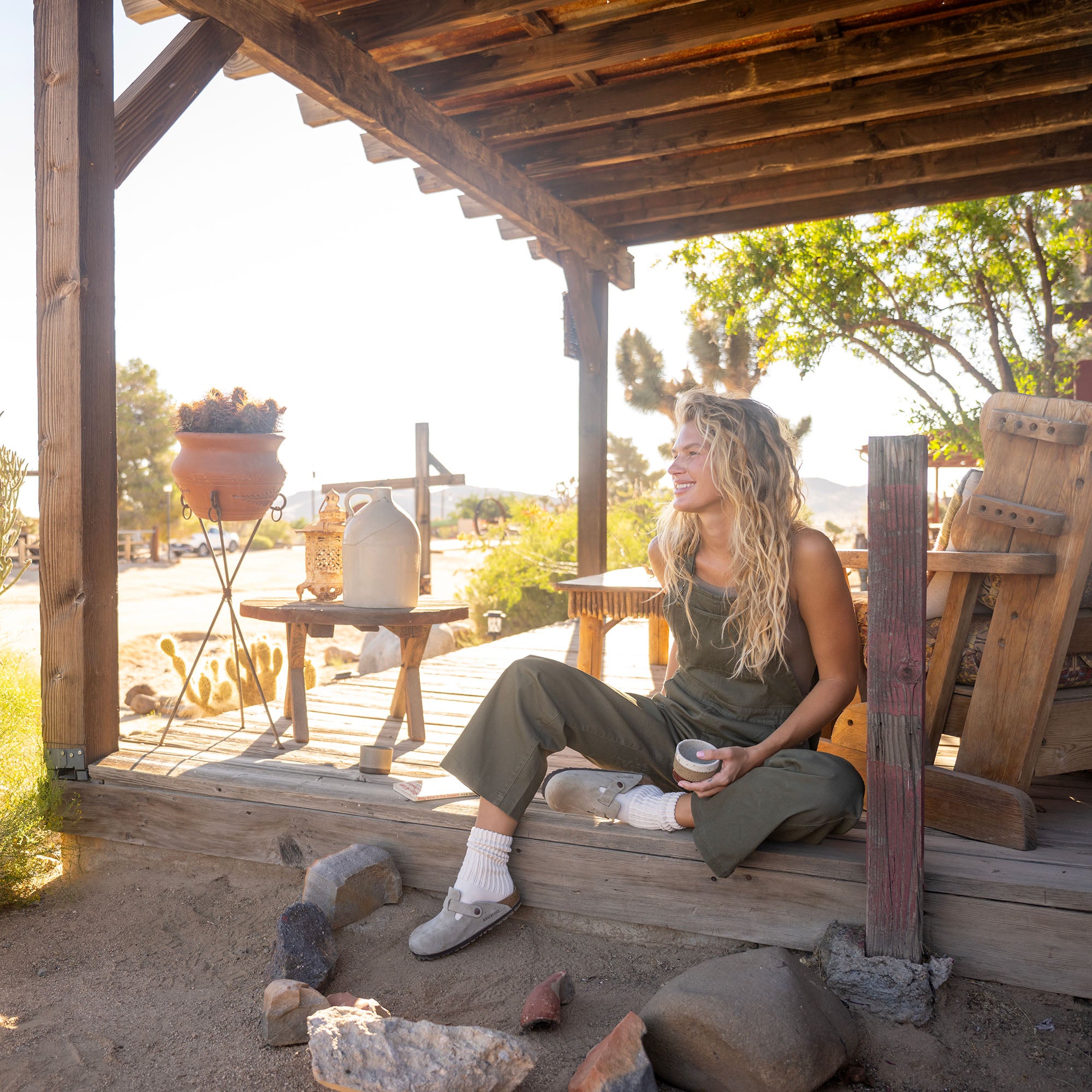 Woman in olive green jumpsuit sitting on wooden pergola deck holding coffee cup in desert landscape setting