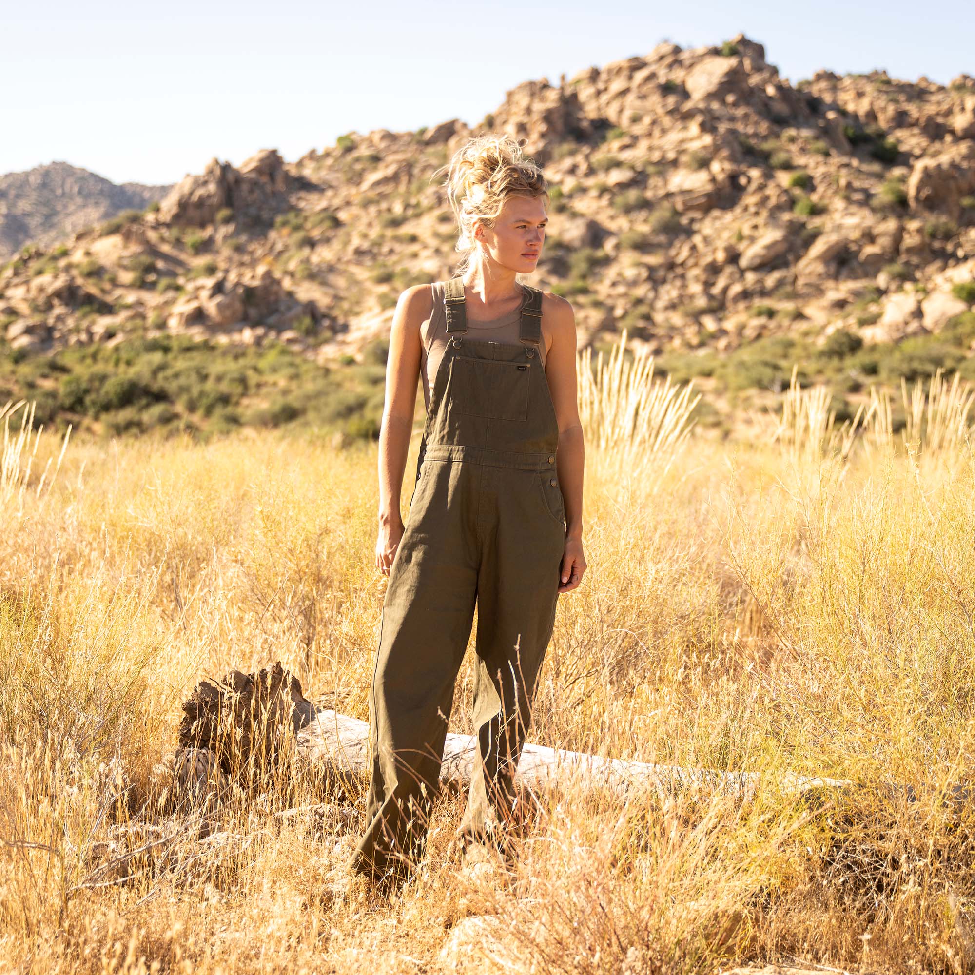 Woman wearing olive-green Costa Jumpsuit standing in desert landscape with rocky hills and dry grassland behind her