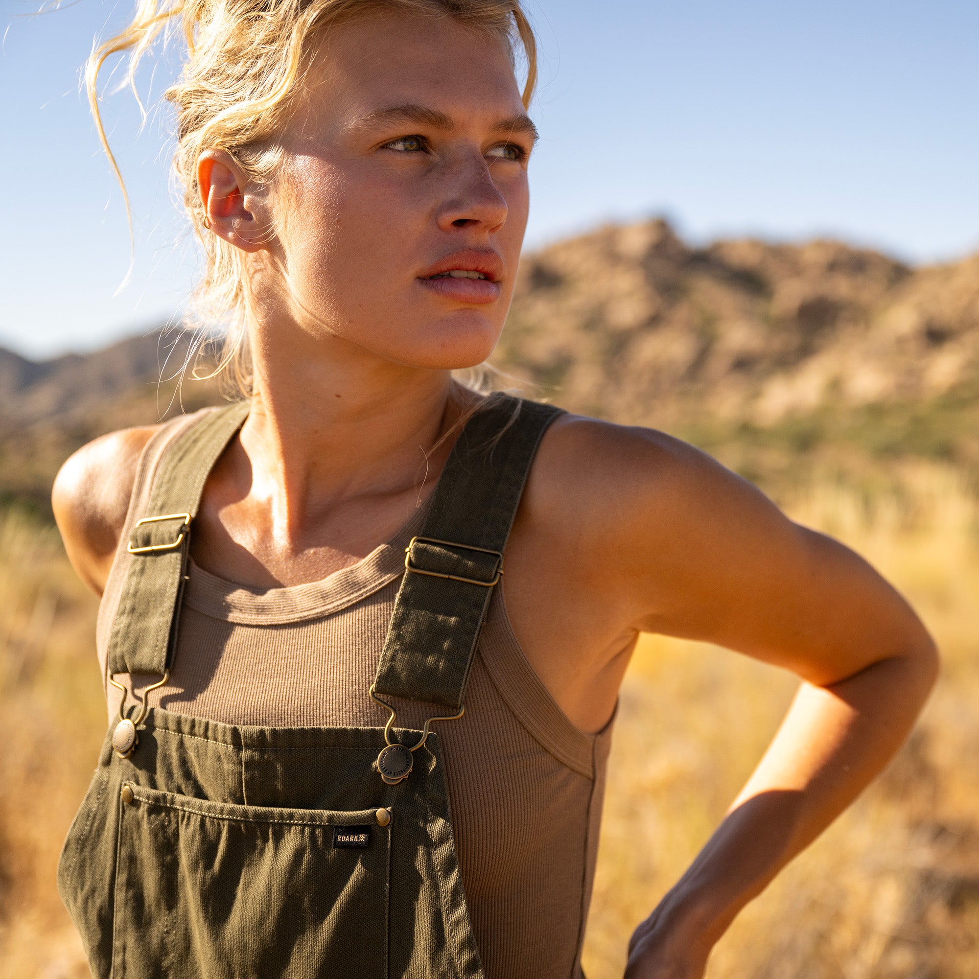 Woman wearing olive green denim jumpsuit over beige ribbed tank top, standing outdoors in desert landscape