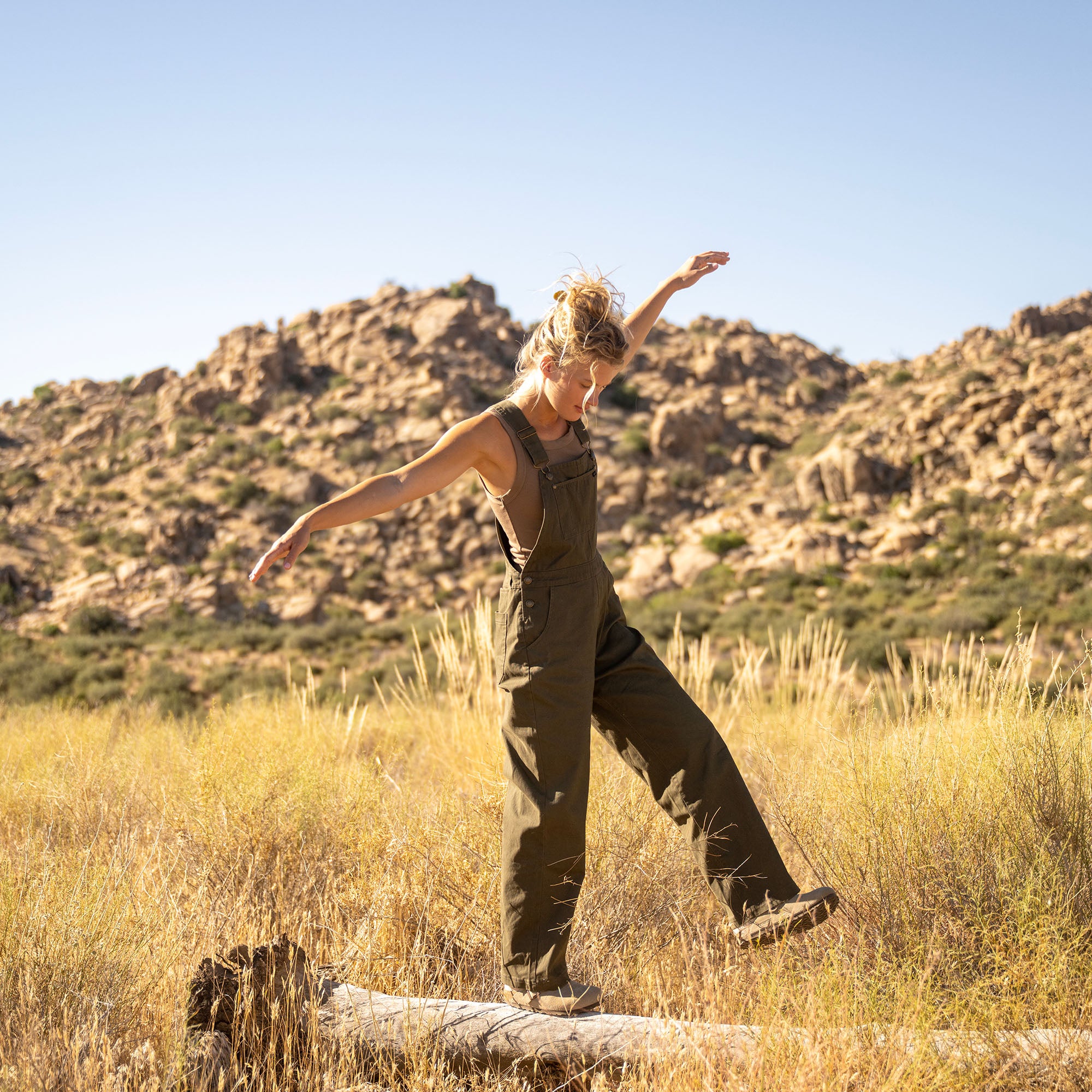 Woman wearing olive green Costa Jumpsuit balancing on rock in desert landscape with rocky hills and dry grass