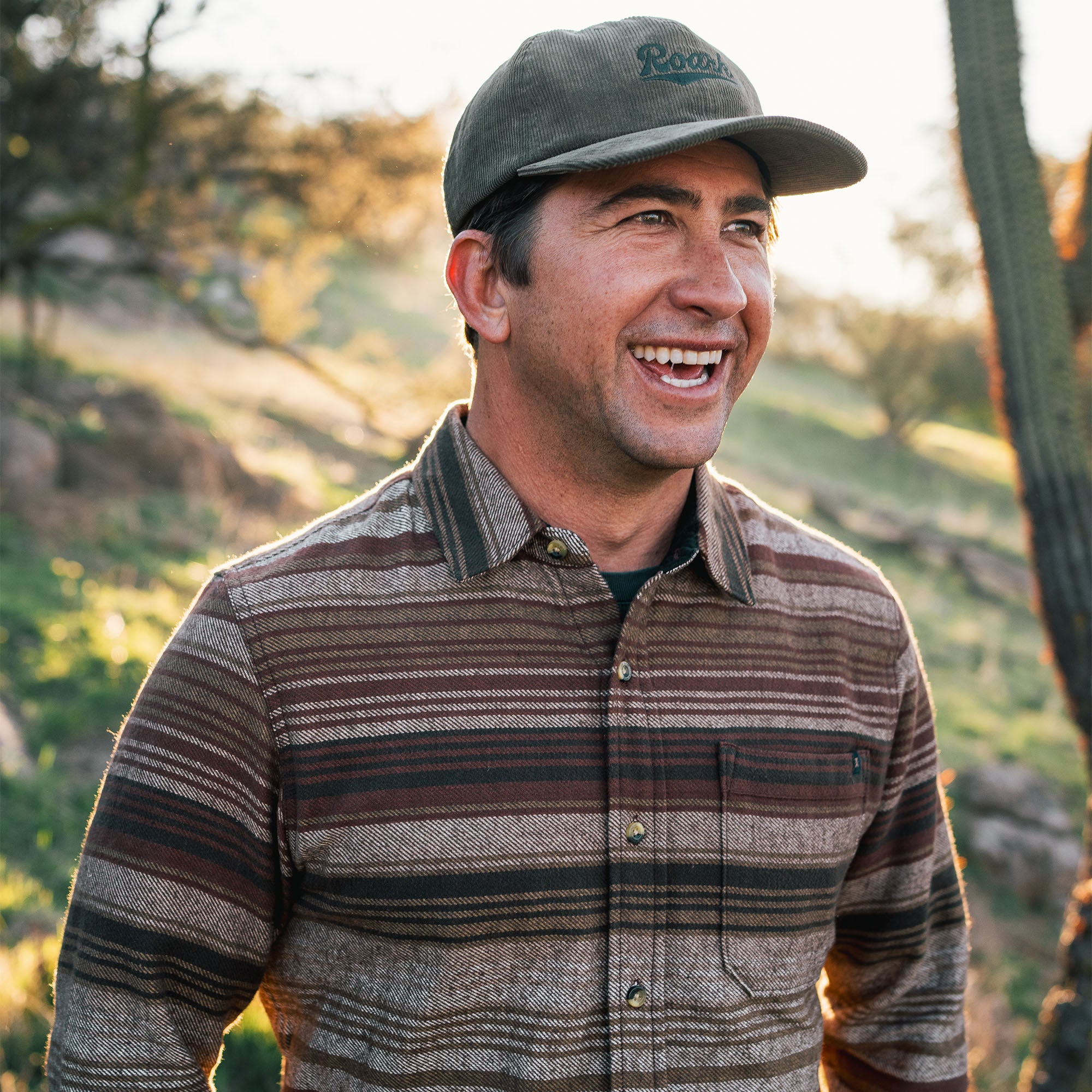 Man smiling in Roark Crossroads Organic Cotton Flannel, paired with a hat, exuding a cheerful outdoor adventure vibe.