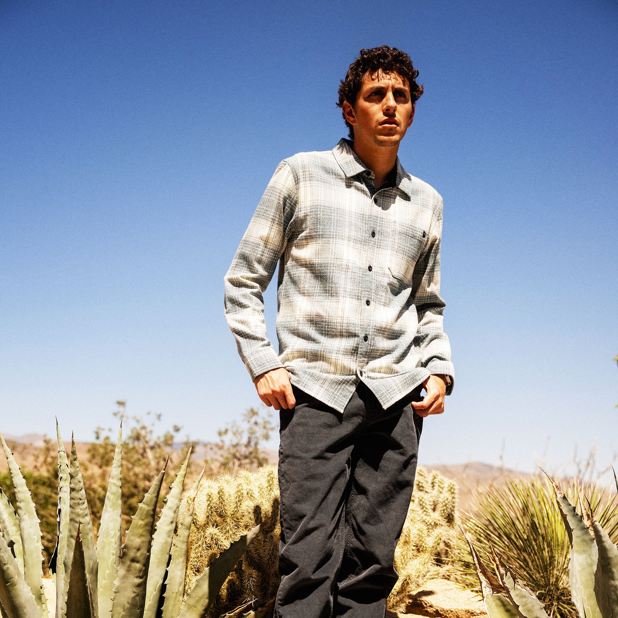 Man wearing cream and gray plaid organic cotton flannel shirt with dark pants standing in desert landscape with cacti