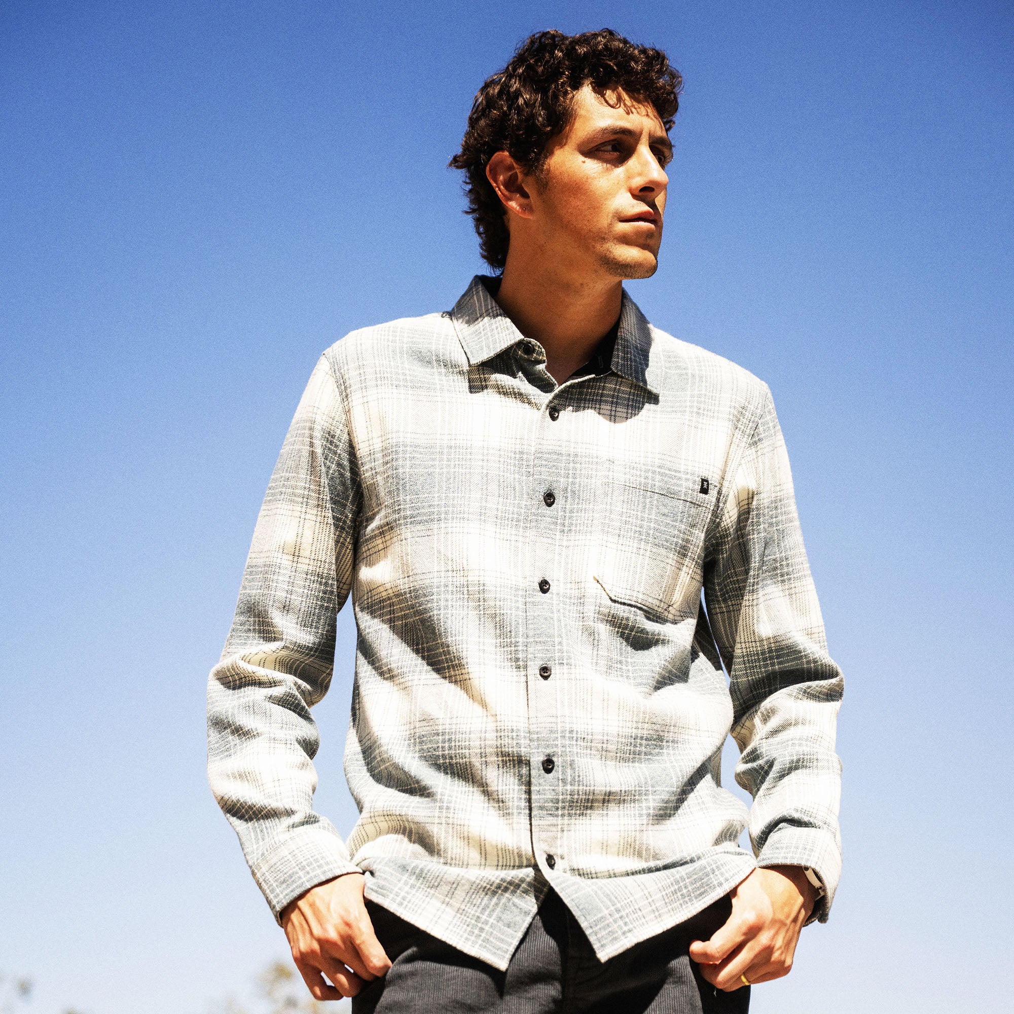 Man wearing cream and gray plaid organic cotton flannel shirt with button front and chest pocket against blue sky