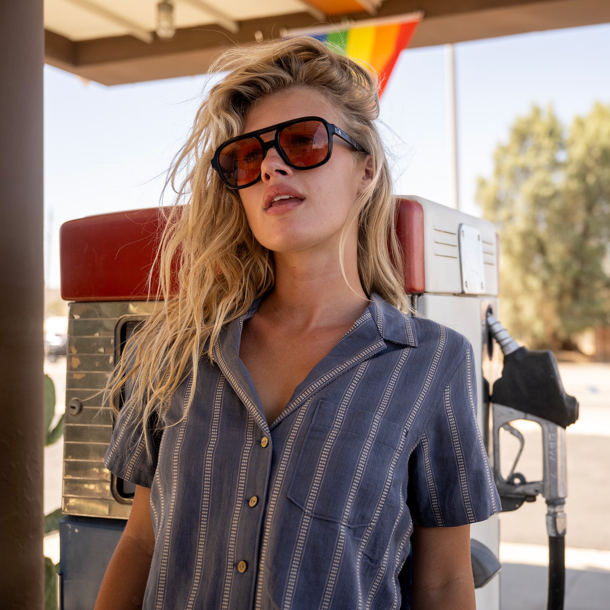 Woman wearing blue pinstriped short-sleeve button-up shirt and black tinted sunglasses standing outdoors under a covered structure