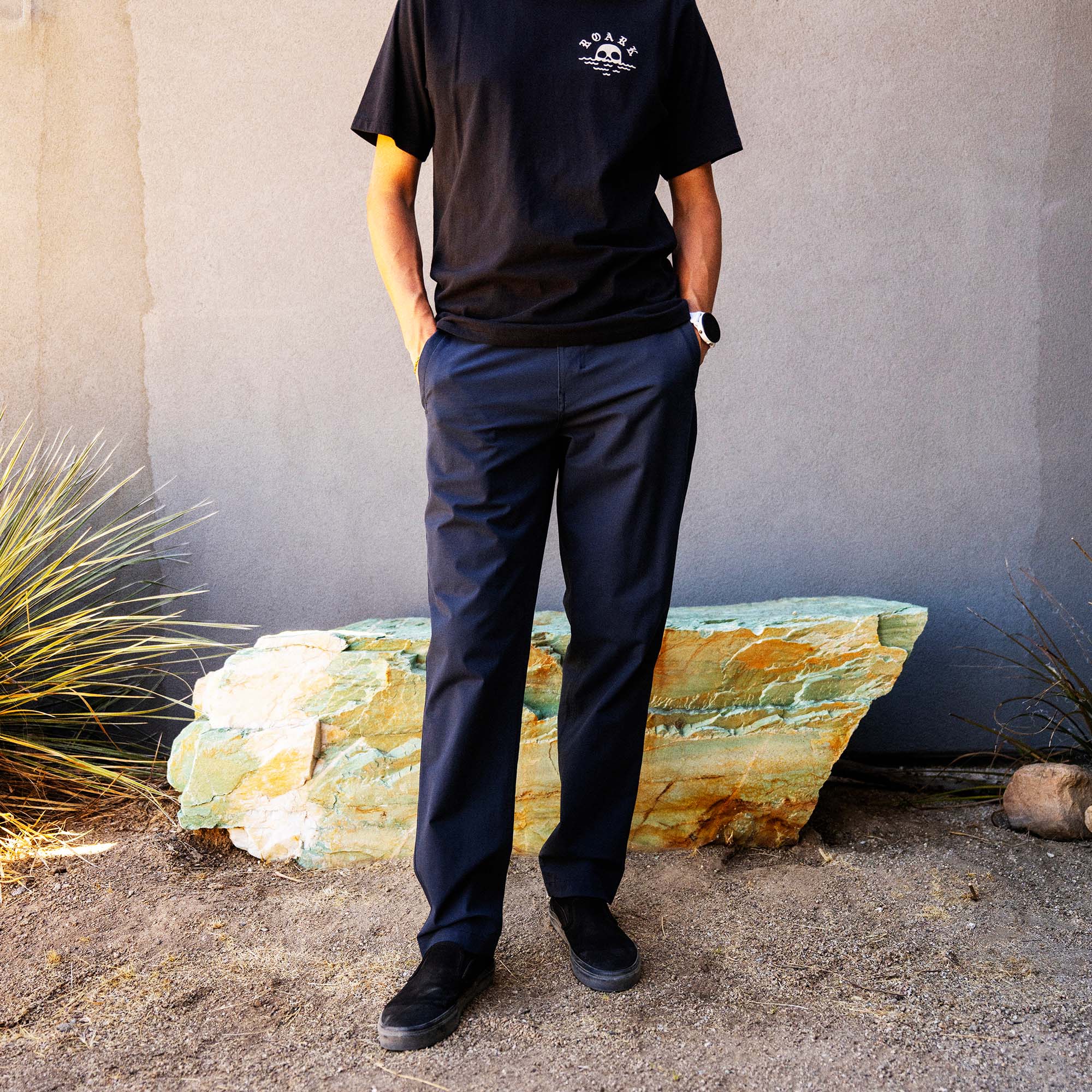 Person wearing black Fear The Sea premium t-shirt and navy chinos standing outdoors against a textured wall