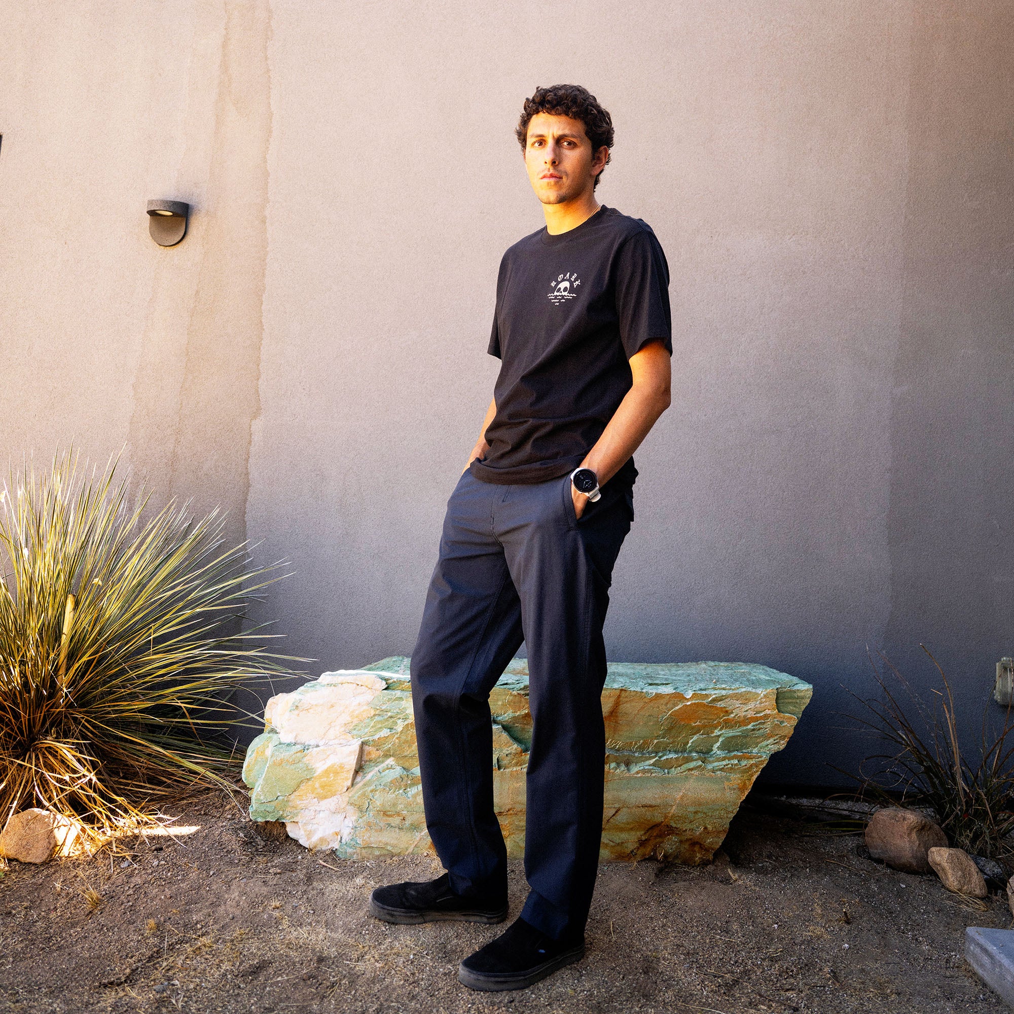 Man wearing black Fear The Sea premium tee with small chest graphic and dark navy trousers standing outdoors against concrete wall