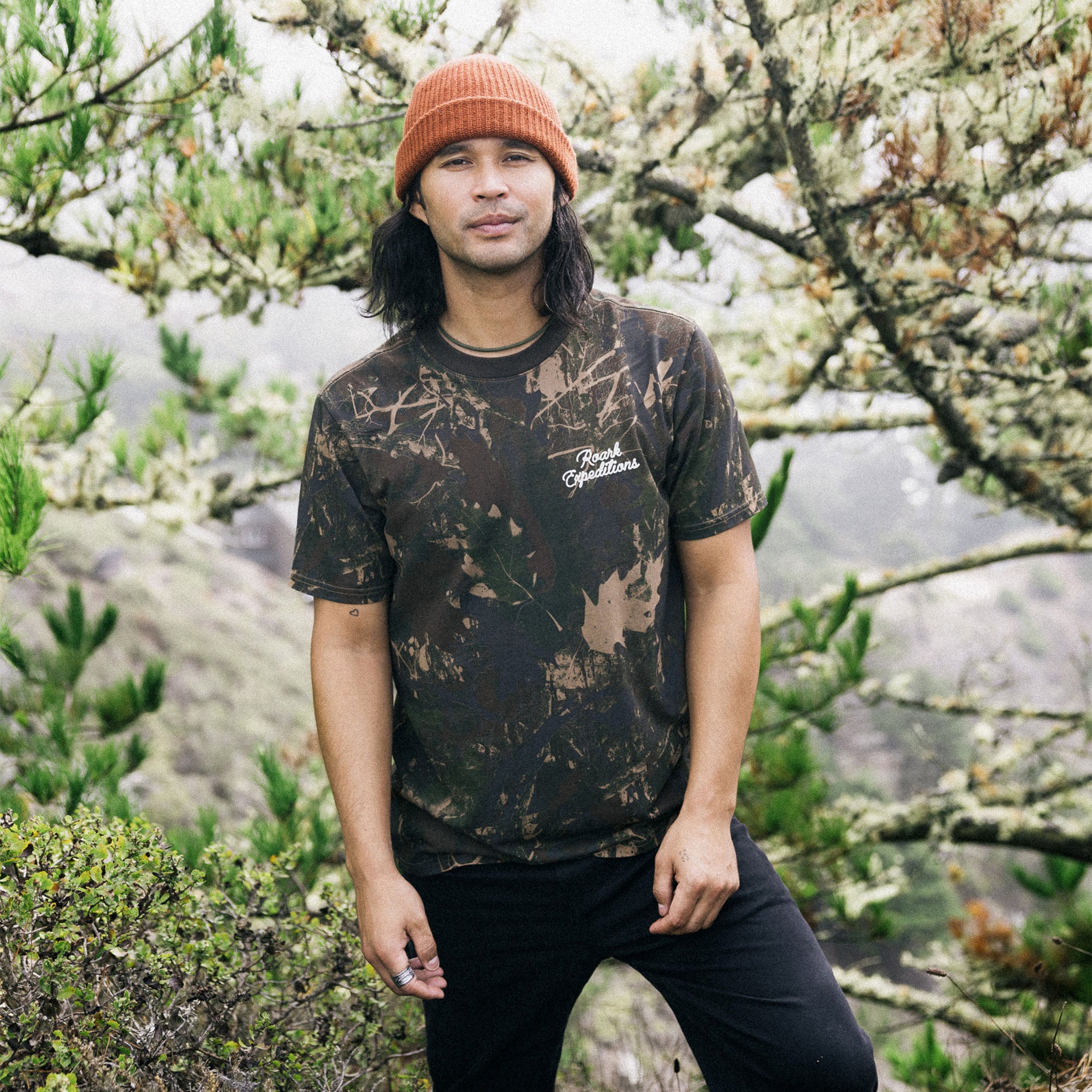 Person wearing a forest camo premium t-shirt by Roark, blending seamlessly with the natural woodland surroundings, emphasizing adventure and outdoor lifestyle.