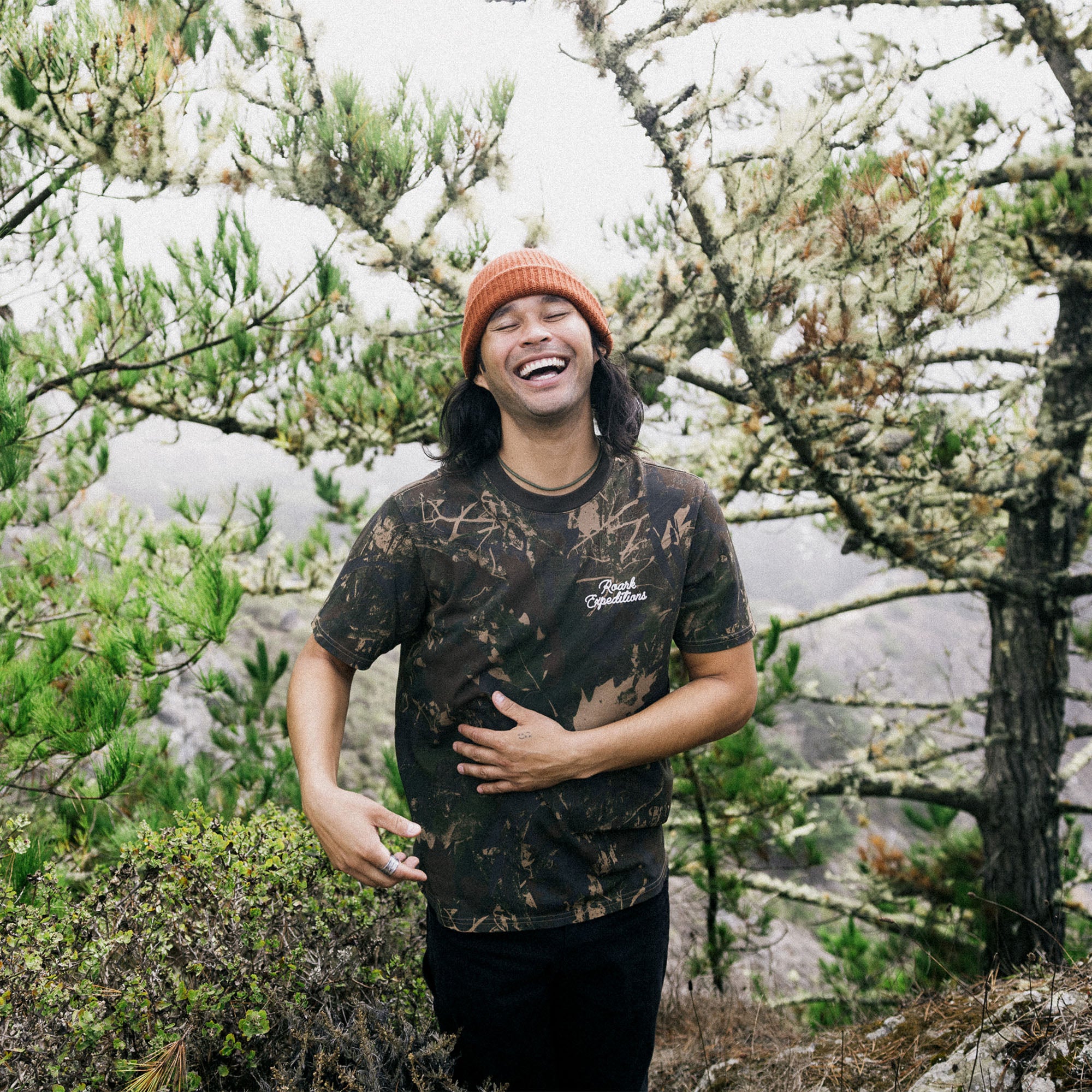 Active person wearing a Roark forest camo premium tee, blending into a natural leafy environment during an outdoor adventure.