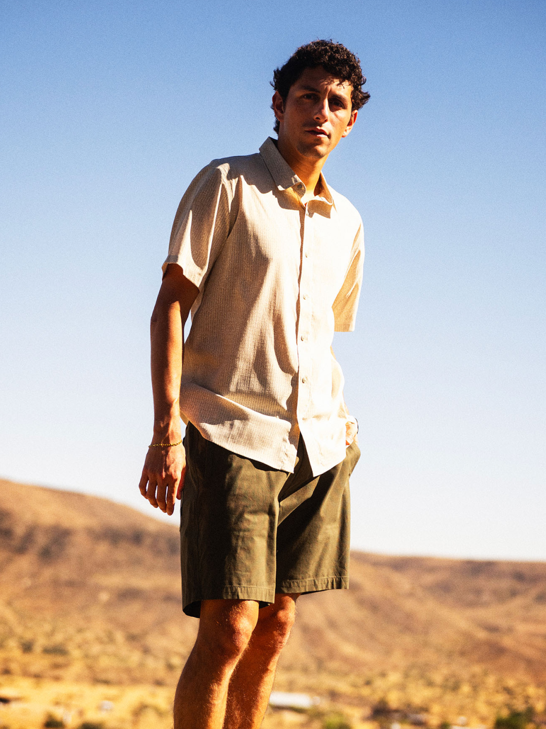Man wearing cream linen short-sleeve button-up shirt and khaki shorts standing in desert landscape under clear sky