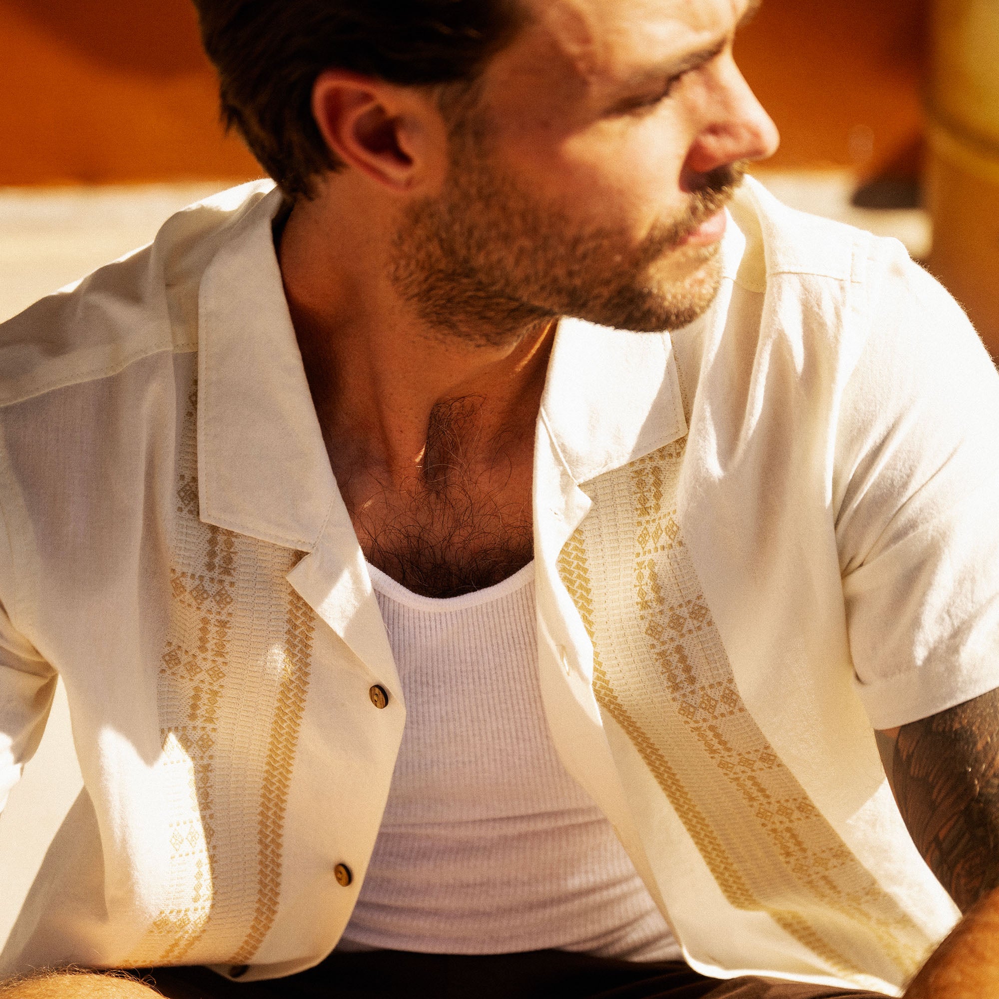 Man wearing cream linen shirt with gold geometric pattern over white ribbed tank top, seated outdoors in warm sunlight