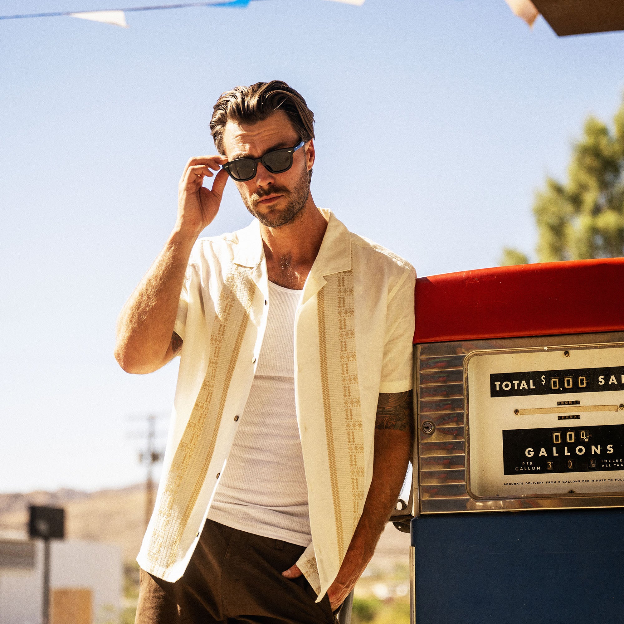 Man wearing cream camp collar shirt and dark sunglasses standing beside vintage gas pump in sunny desert setting