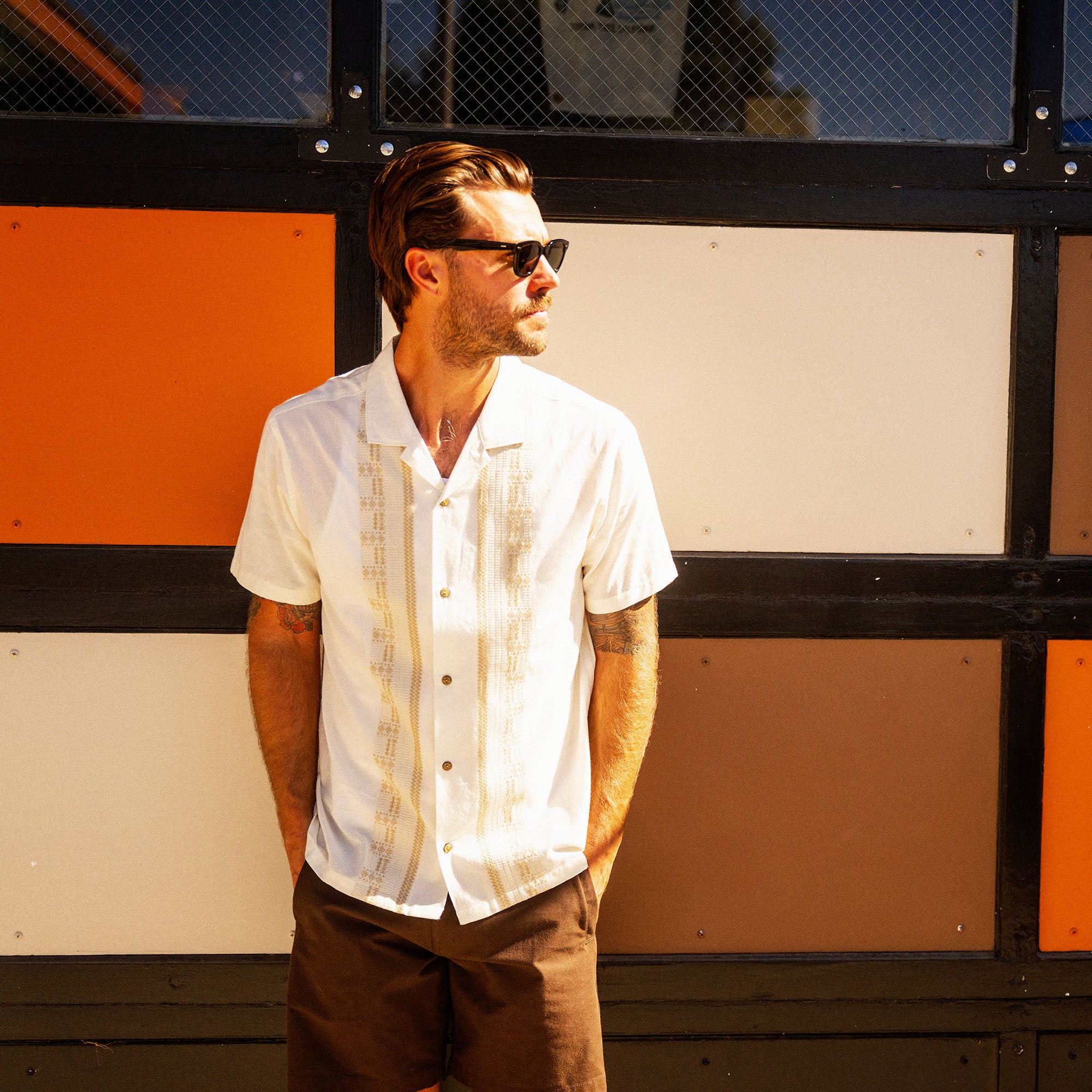 Man wearing white short-sleeve button-up shirt with tan textured stripes, paired with brown shorts and dark sunglasses