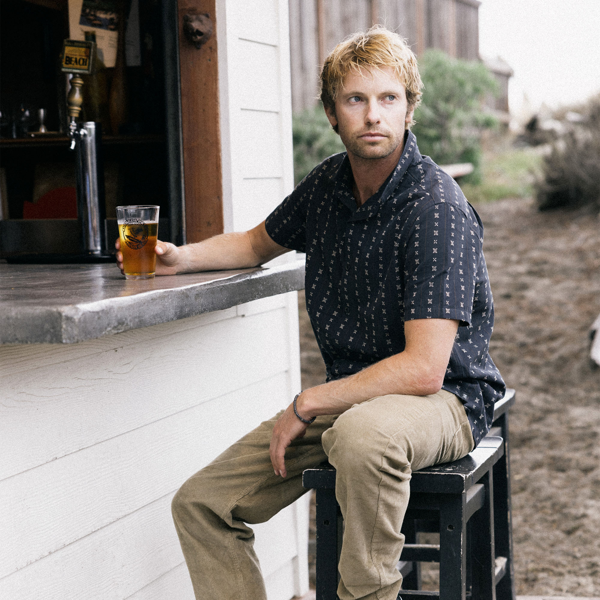 Man with facial hair sitting in a rugged outdoor setting, wearing Roark's denim Gonzo dress shirt with rolled-up sleeves, embodying an adventurous spirit.