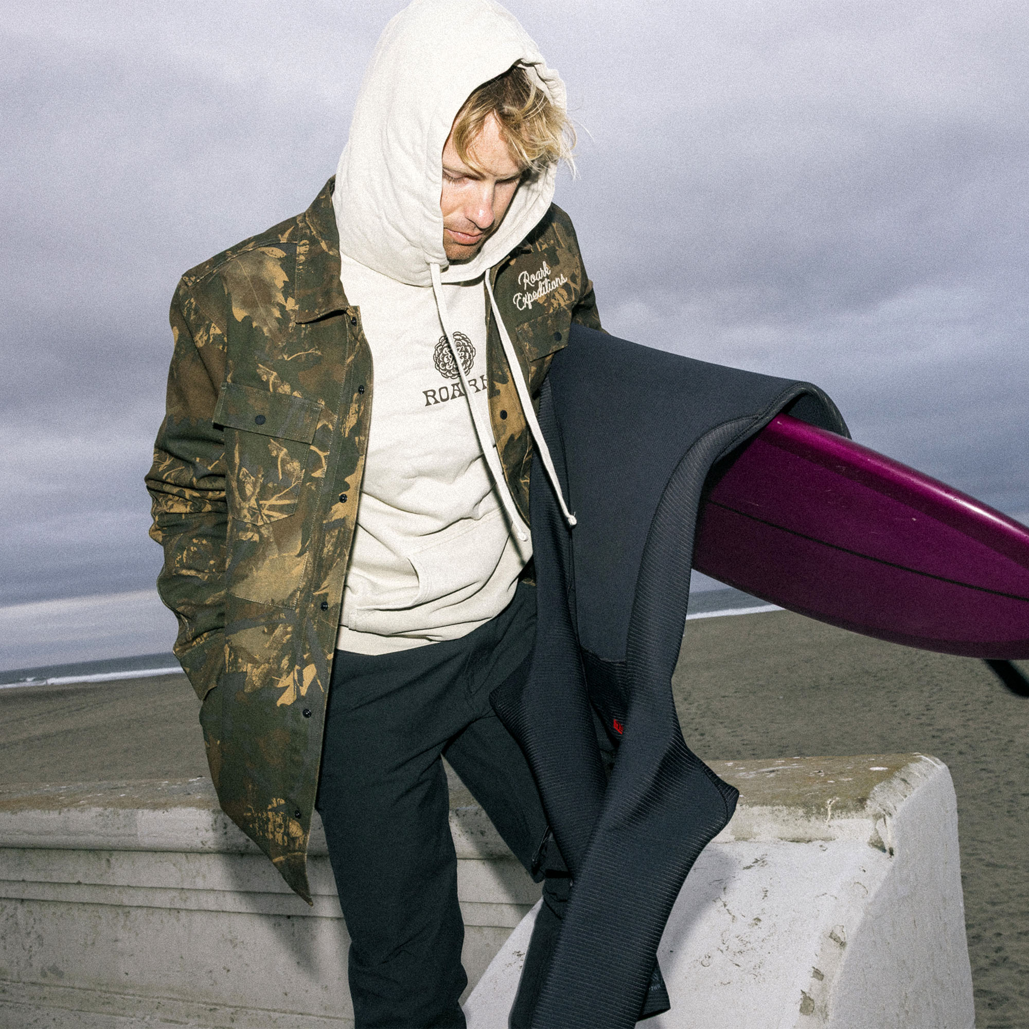 Person wearing Roark's Hebrides Unlined Jacket in a camouflage pattern, exploring a natural, outdoor setting with hood up and sleeves visible.