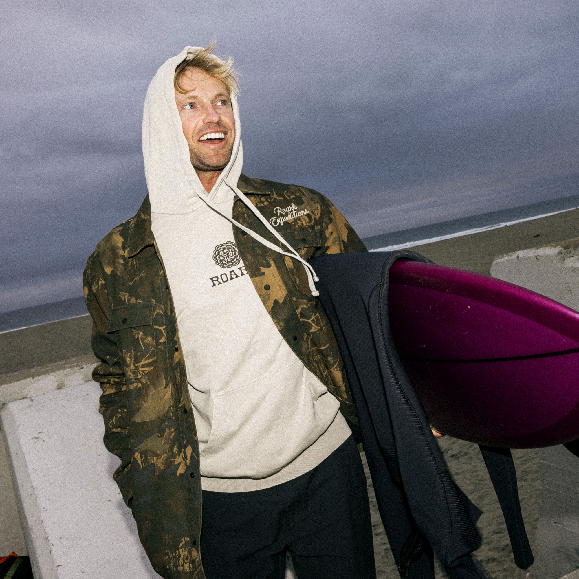 Surfer with a surfboard wearing the Hebrides Unlined Jacket by Roark, standing by the sea with rolling ocean waves in the background, illustrating the adventurous spirit of Roark's outdoor apparel.