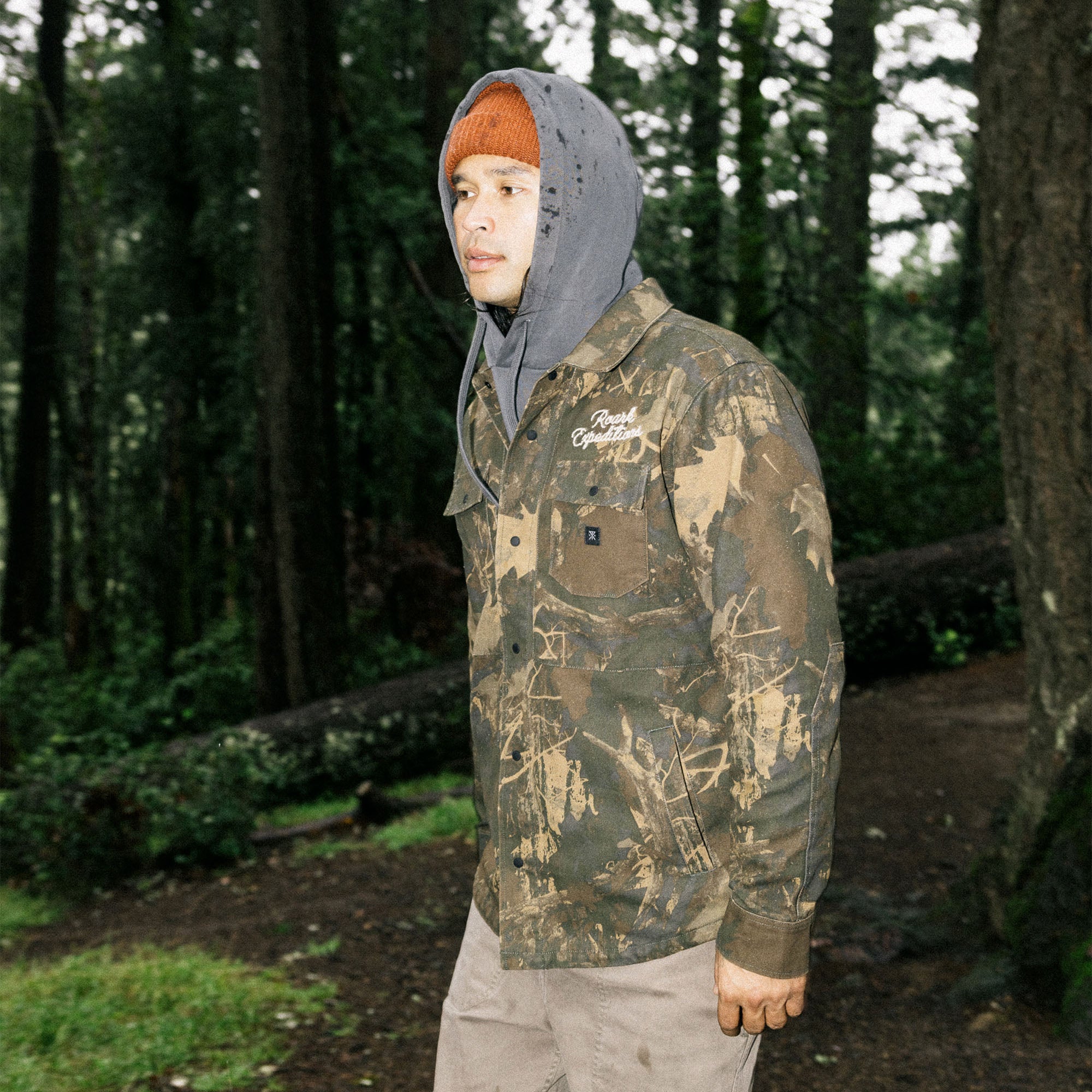 Roark Hebrides unlined woodland camouflage jacket with hood displayed in a natural outdoor setting, emphasizing adventure and durability.